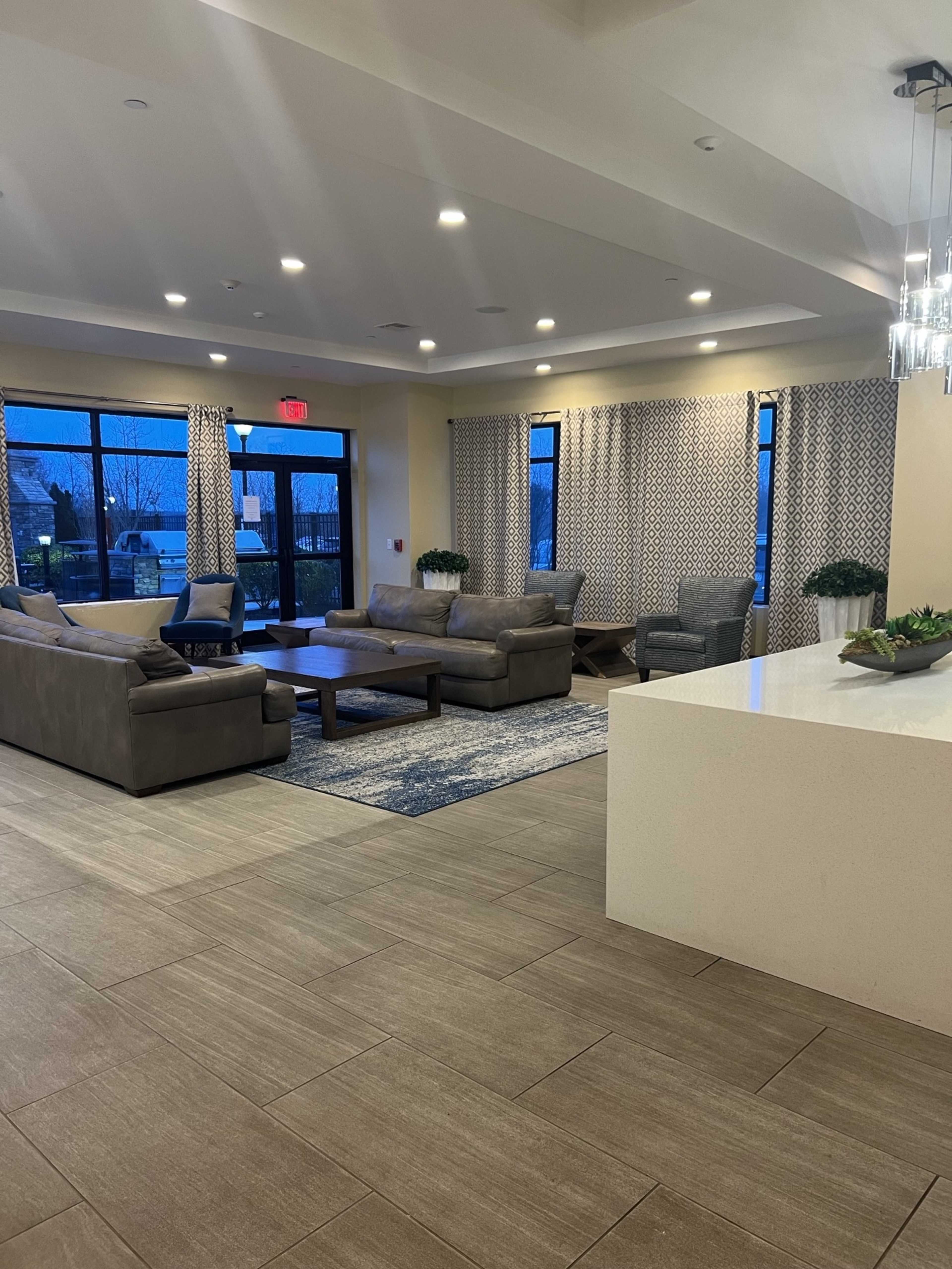 A modern lounge area featuring gray sofas, a coffee table, and large windows with patterned curtains, set on a tile floor.