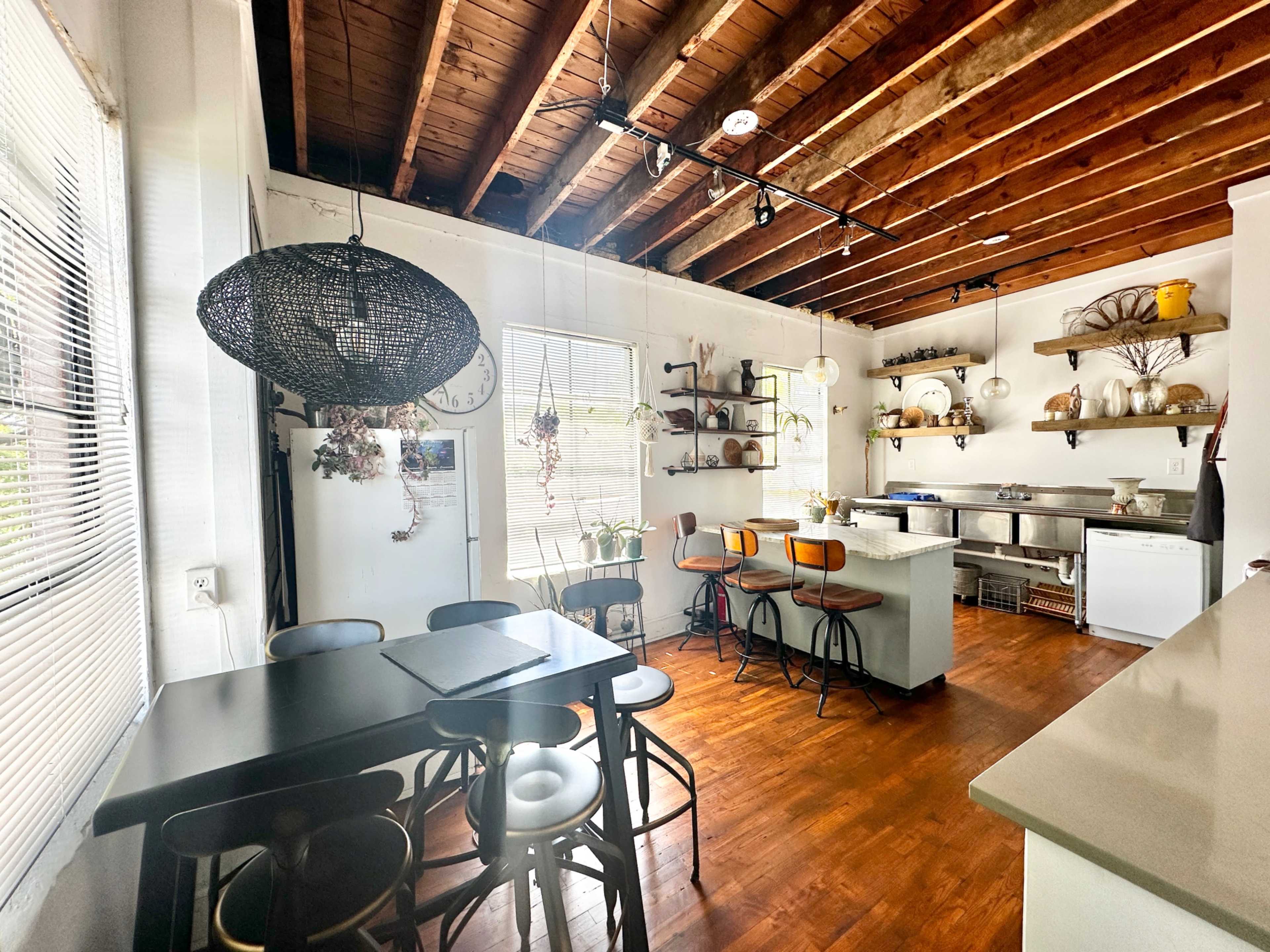 A kitchen with exposed wooden beams, a dining table with black chairs, and a breakfast bar with stools, along with various kitchen appliances and shelves displaying dishes.