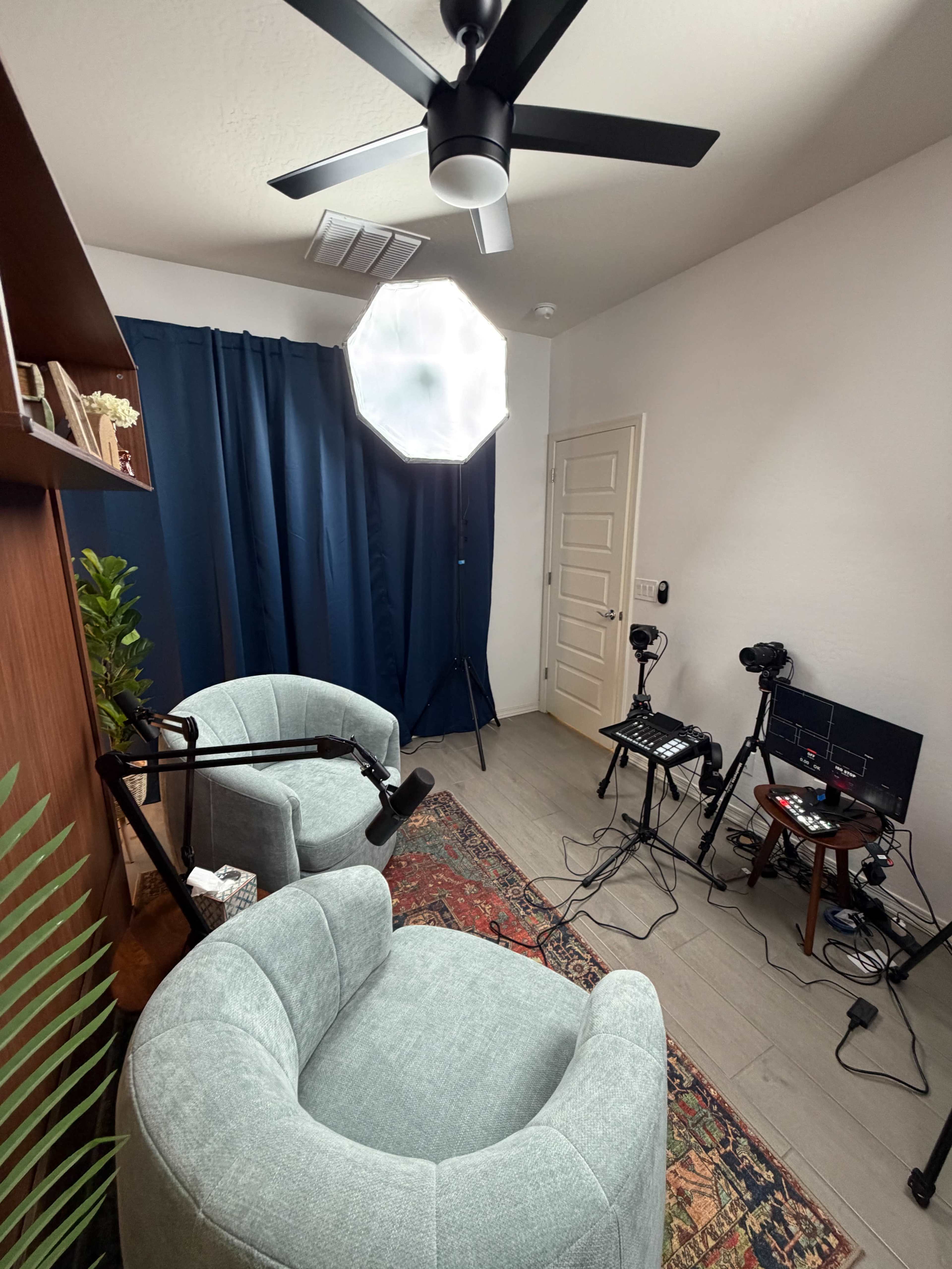 The image shows a small room with two upholstered chairs, a large softbox lighting setup, a table with multiple cameras and equipment, and a curtain in the background.