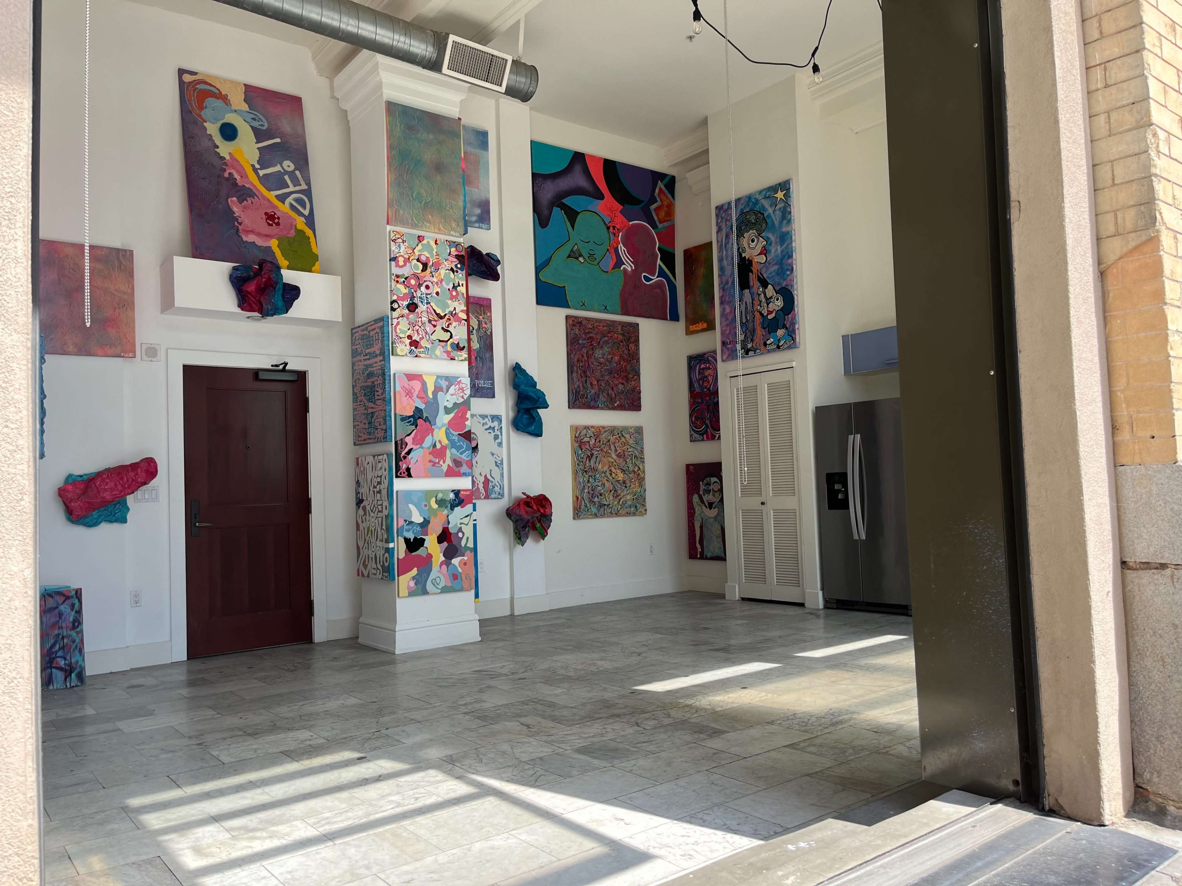 The image shows an art gallery interior featuring colorful abstract paintings on the walls and a stainless steel refrigerator in the corner.