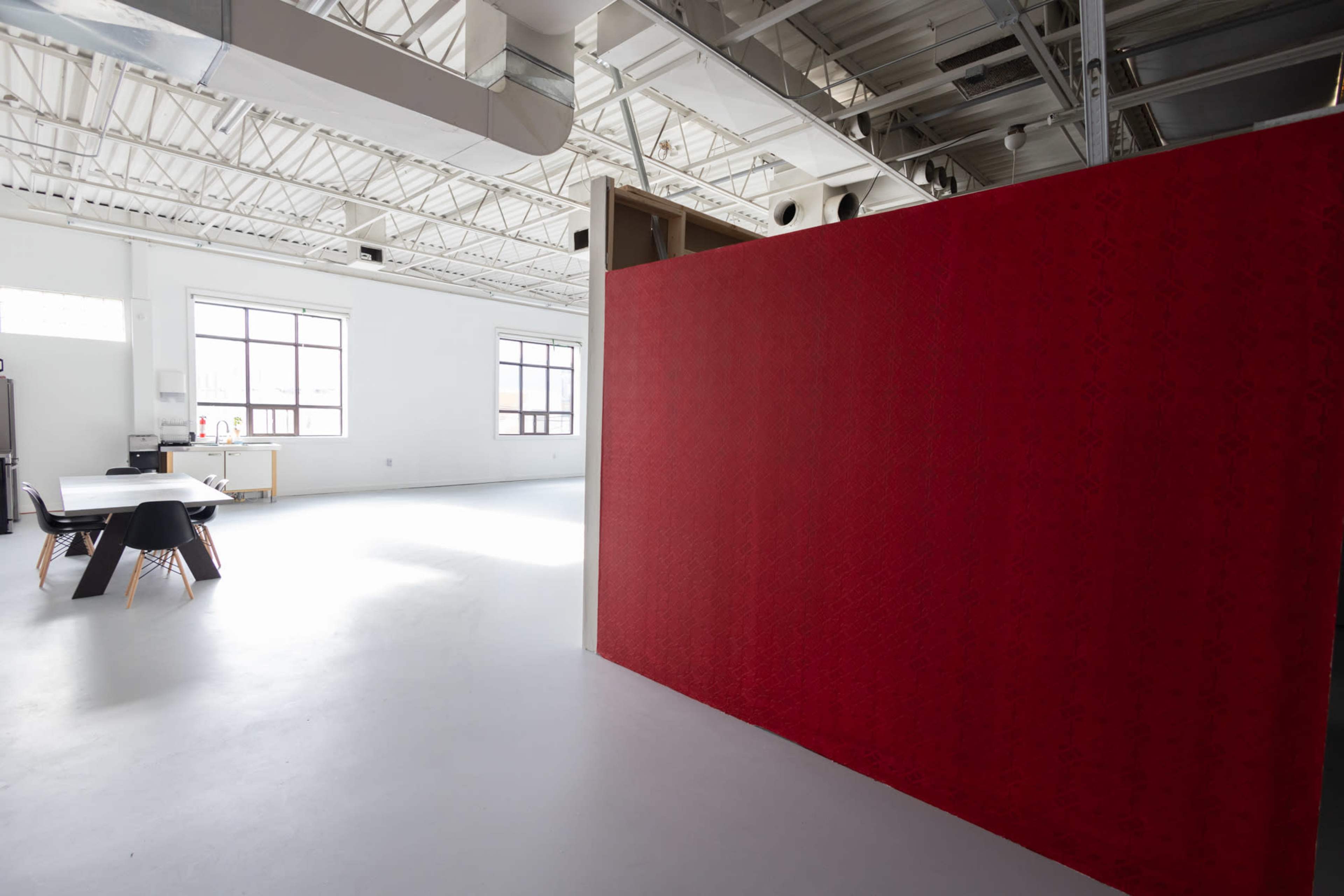 A large, bright room with high ceilings features a red partition and a table with chairs in the background.
