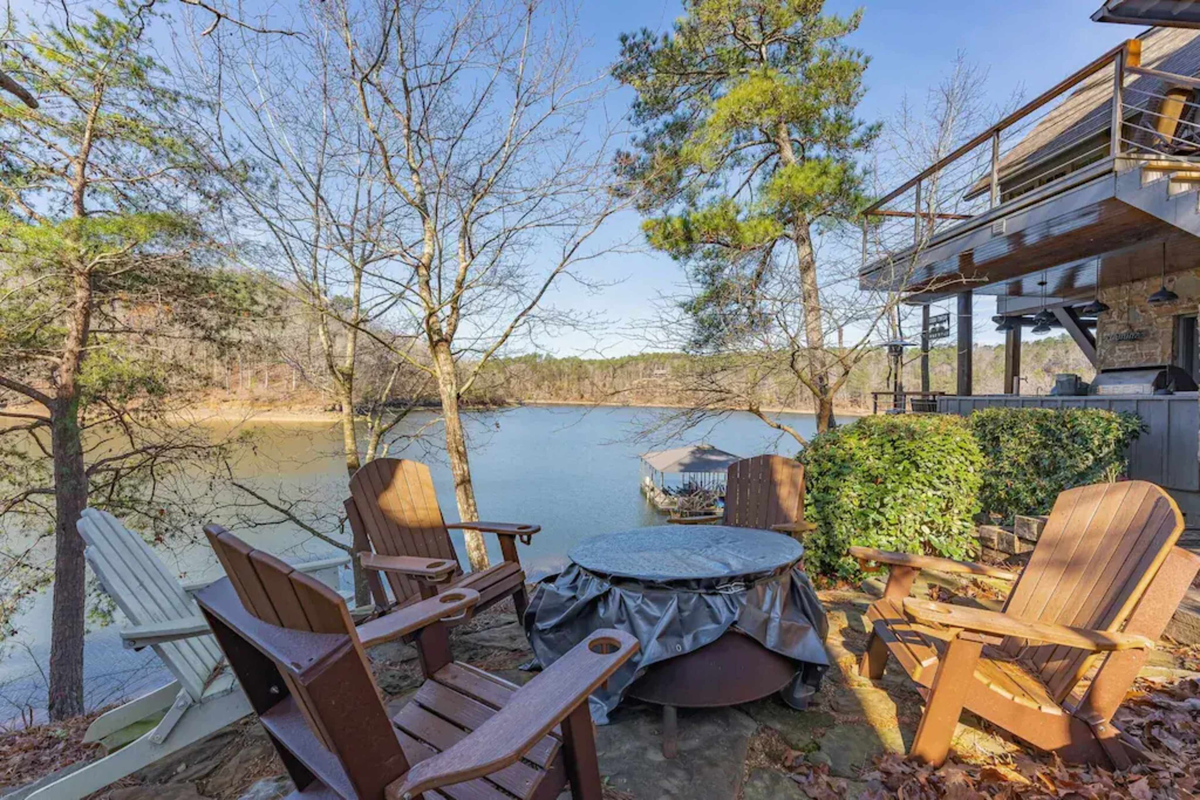 A seating area with wooden chairs surrounds a fire pit, overlooking a lake surrounded by trees.