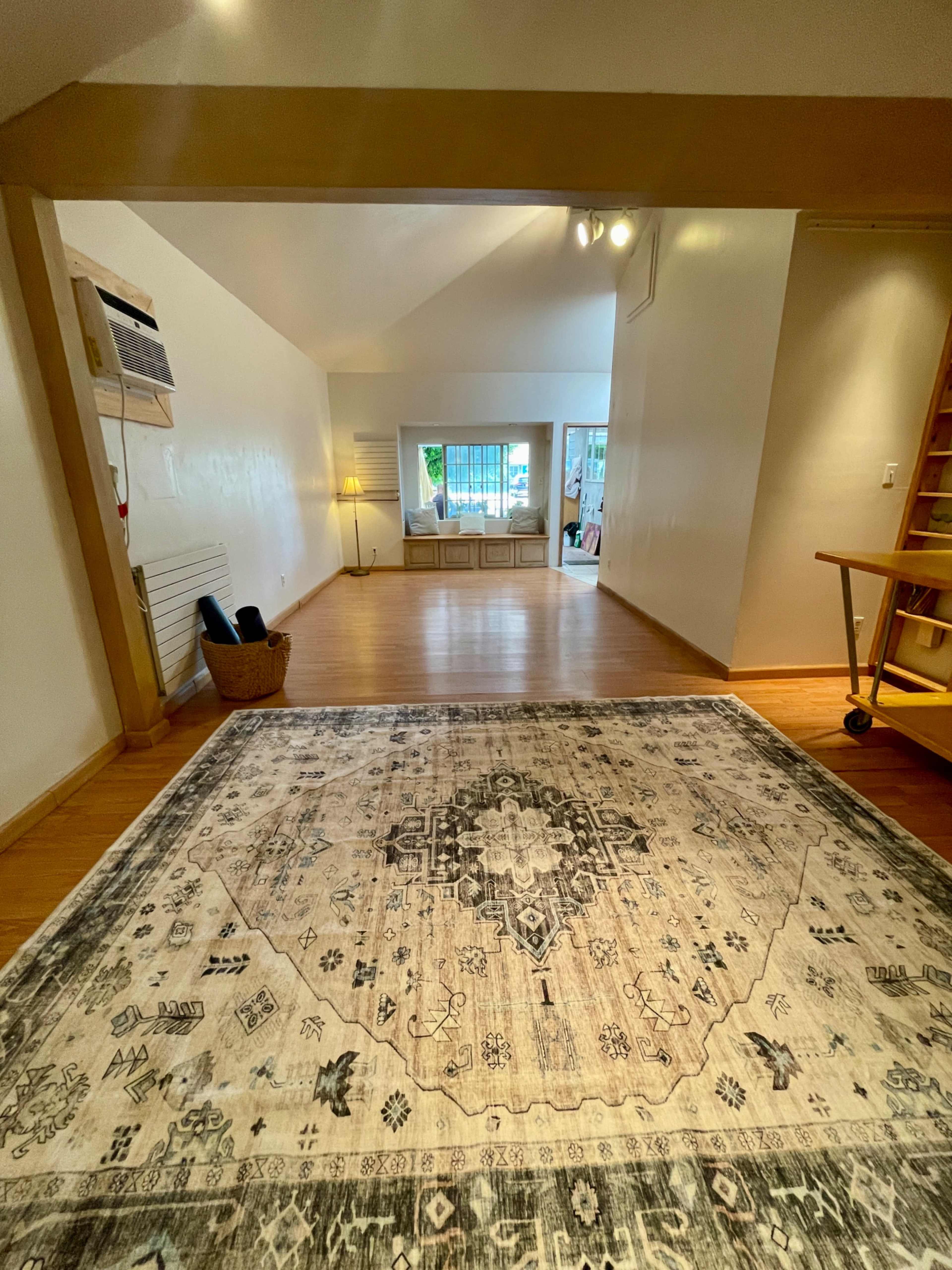 The image shows a spacious, well-lit room featuring a large decorative rug, with a staircase on the right and a bright area visible in the background.
