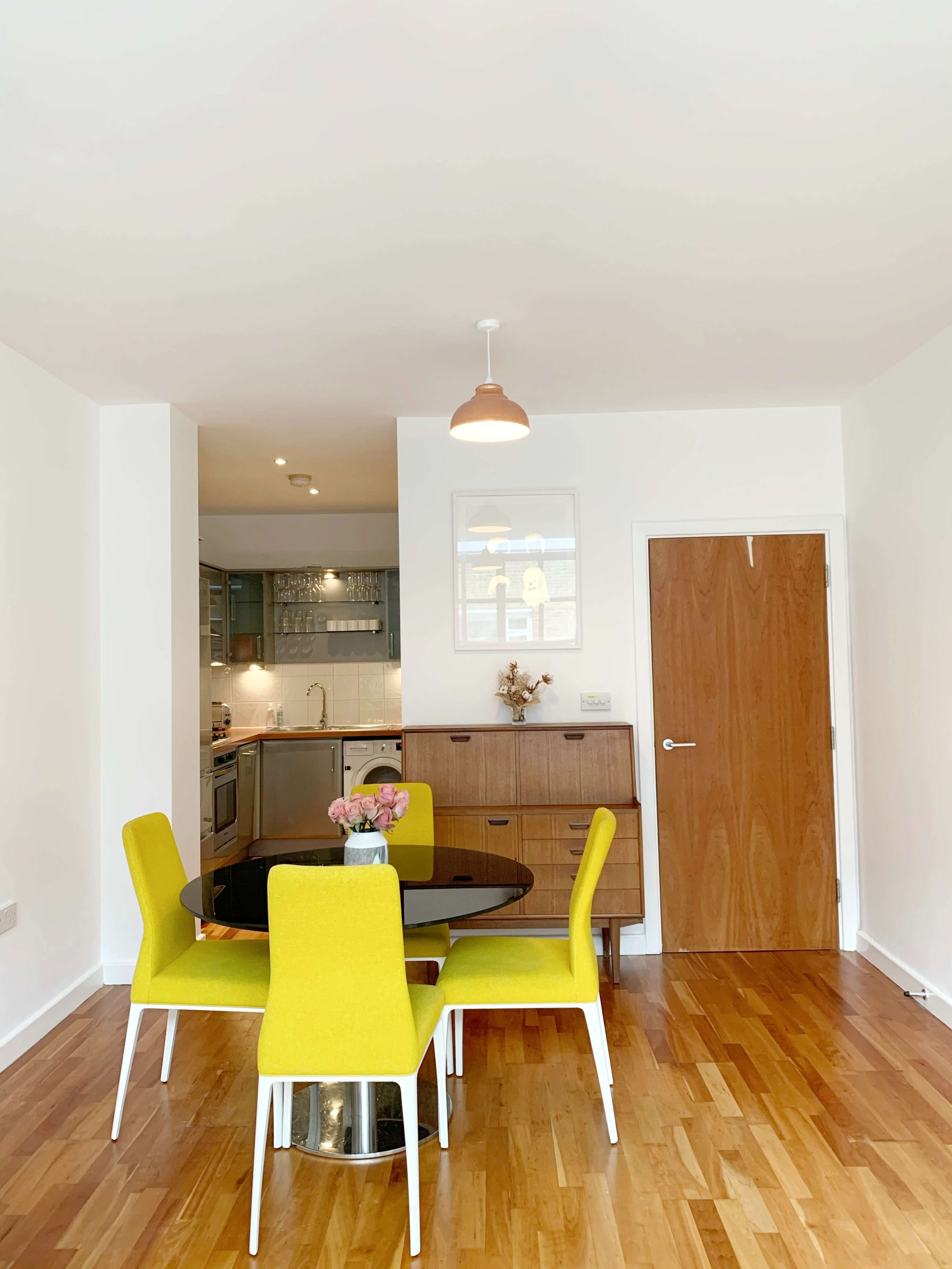 A dining area features a round table surrounded by four yellow chairs, with a wooden cabinet and a door leading to another room in the background.