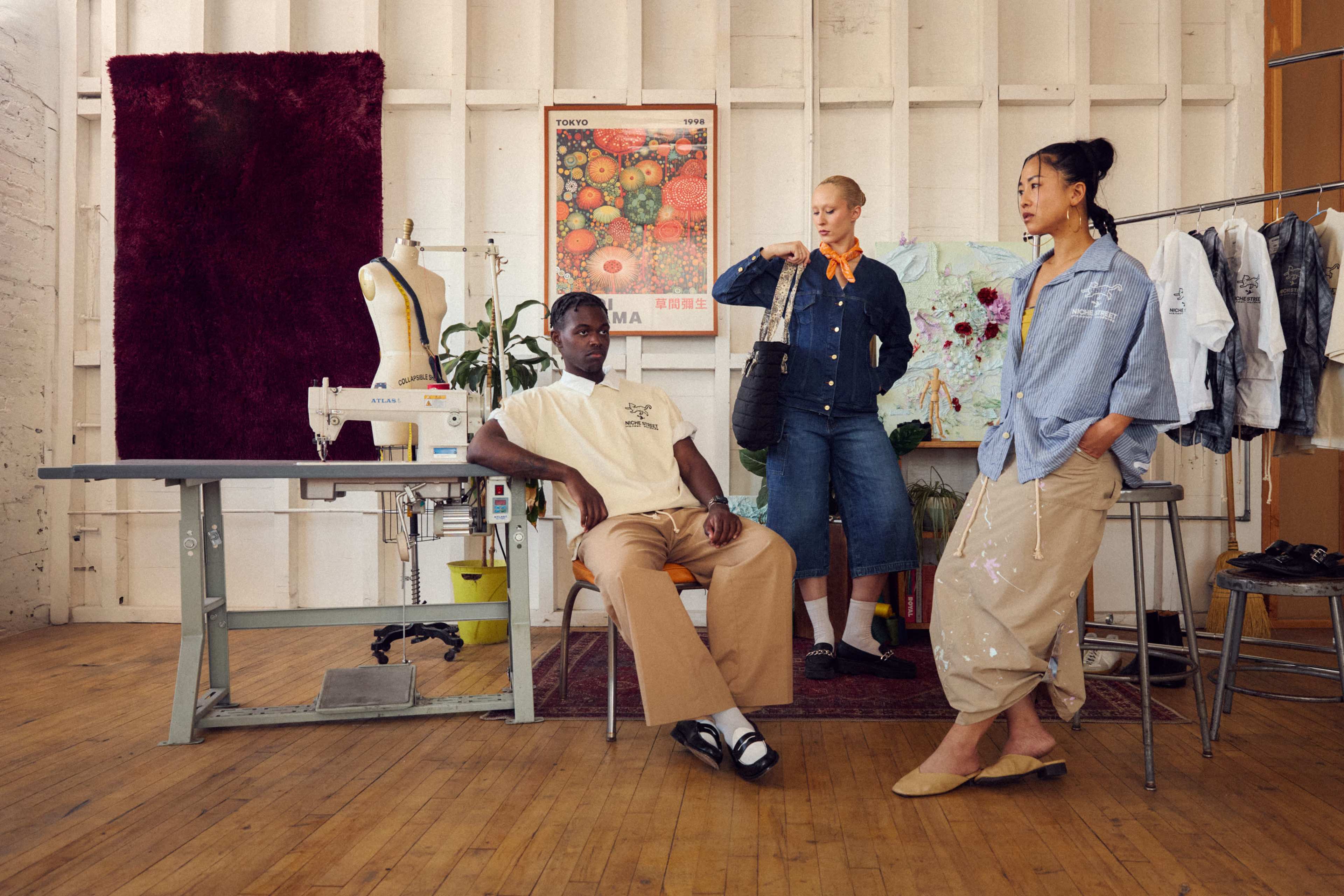 Three individuals are posed in an artistically decorated studio, with one seated at a table and the others standing nearby, surrounded by sewing equipment and colorful artwork.