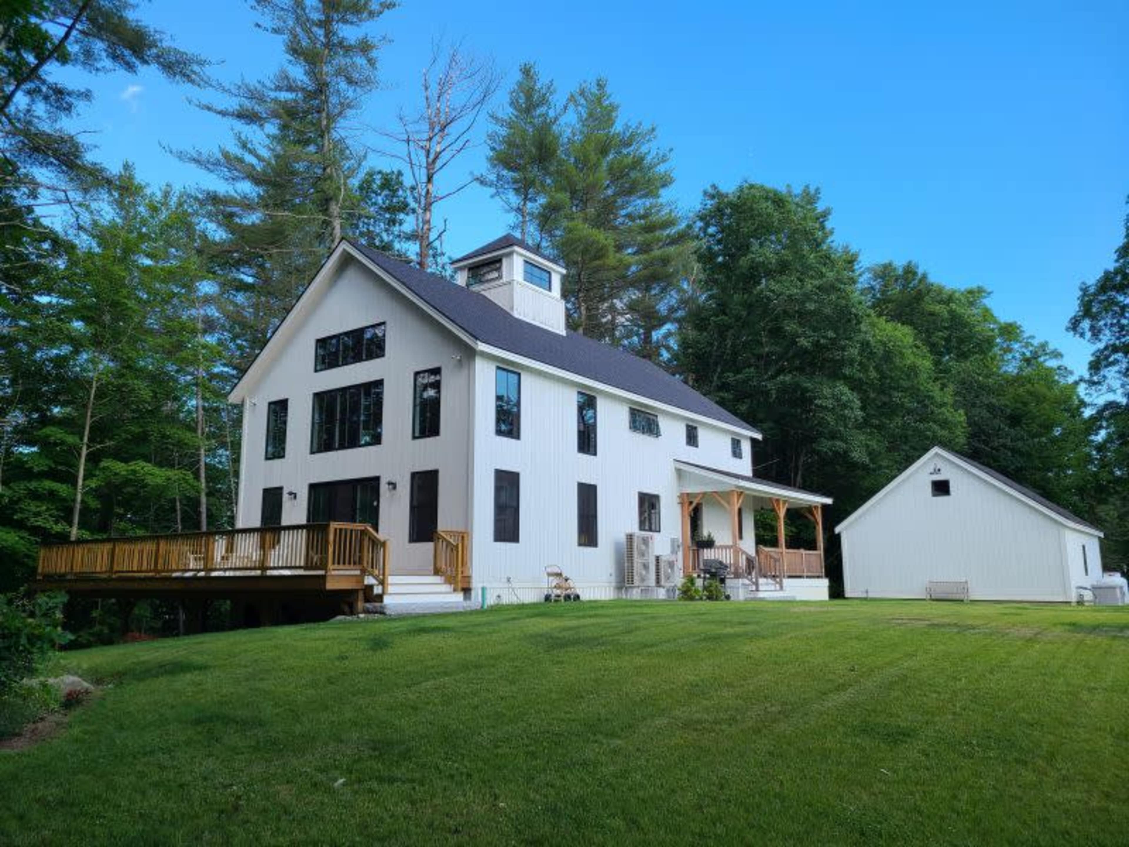 A modern two-story house with a large deck and an adjacent outbuilding, surrounded by tall trees and a grassy lawn.