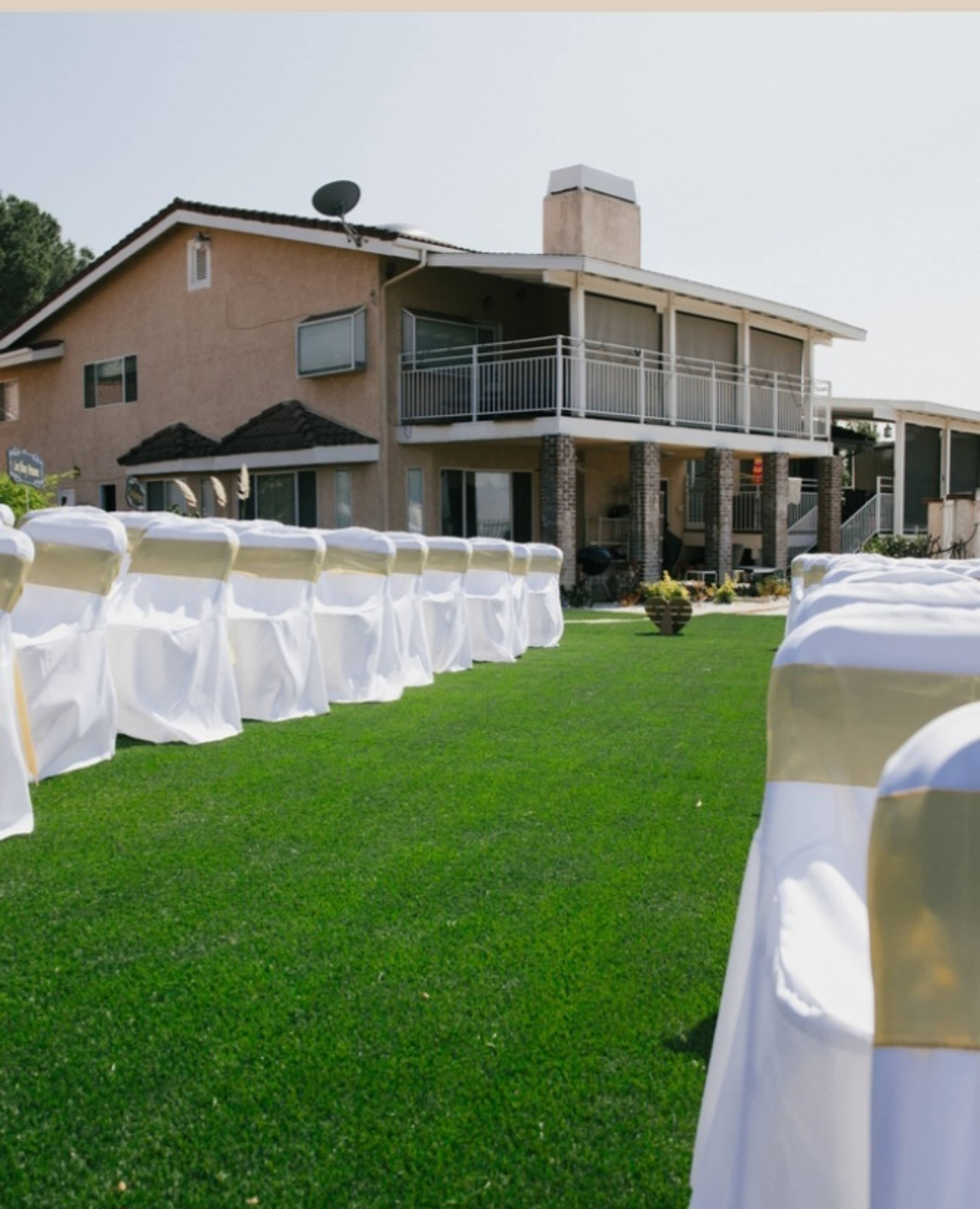 A neatly arranged outdoor wedding setup with white chairs adorned with gold sashes, positioned on a green lawn in front of a two-story house.