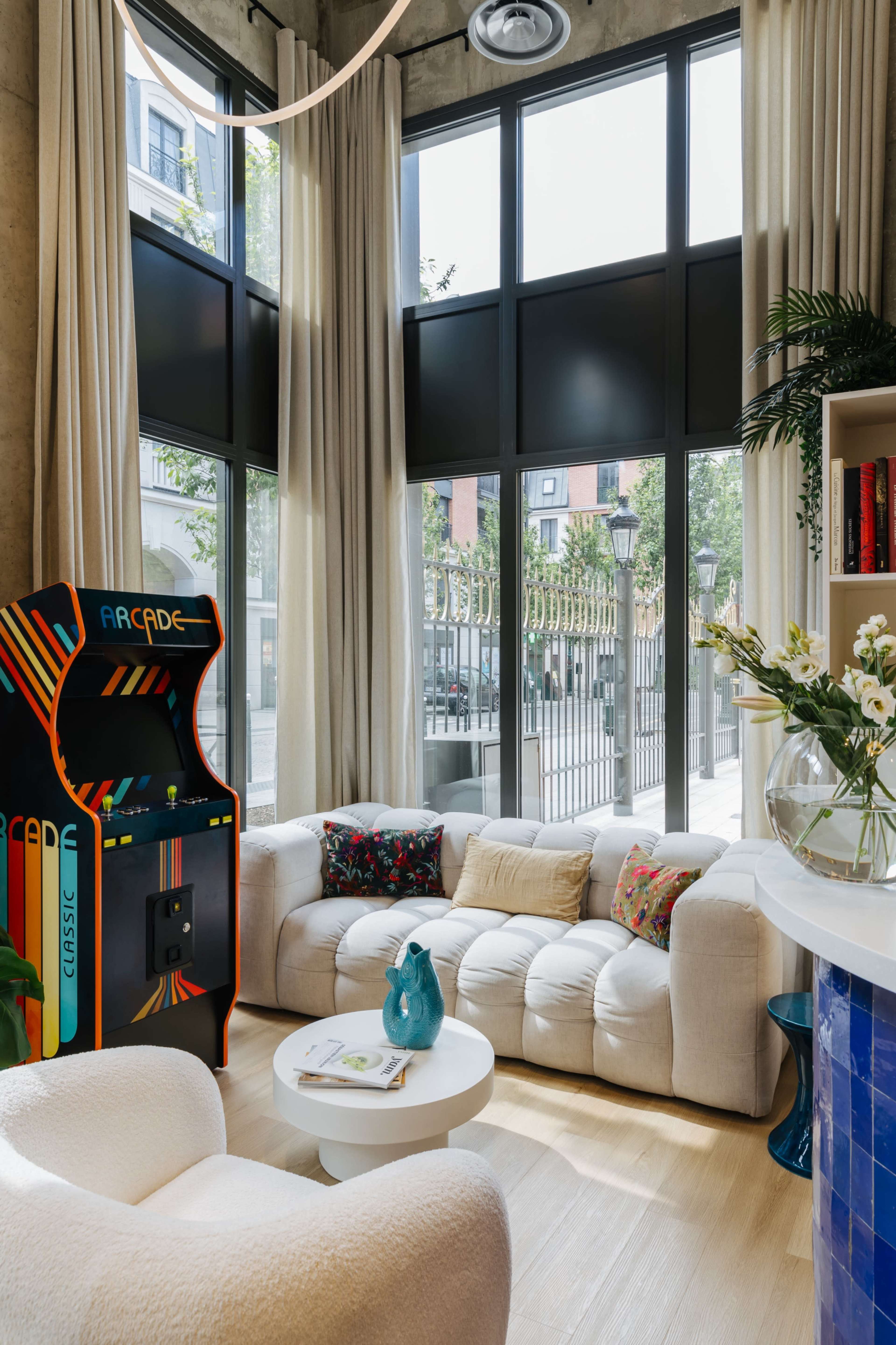 A modern living room features a curved sofa, an arcade machine, and large windows with sheer curtains, allowing natural light to enter the space.