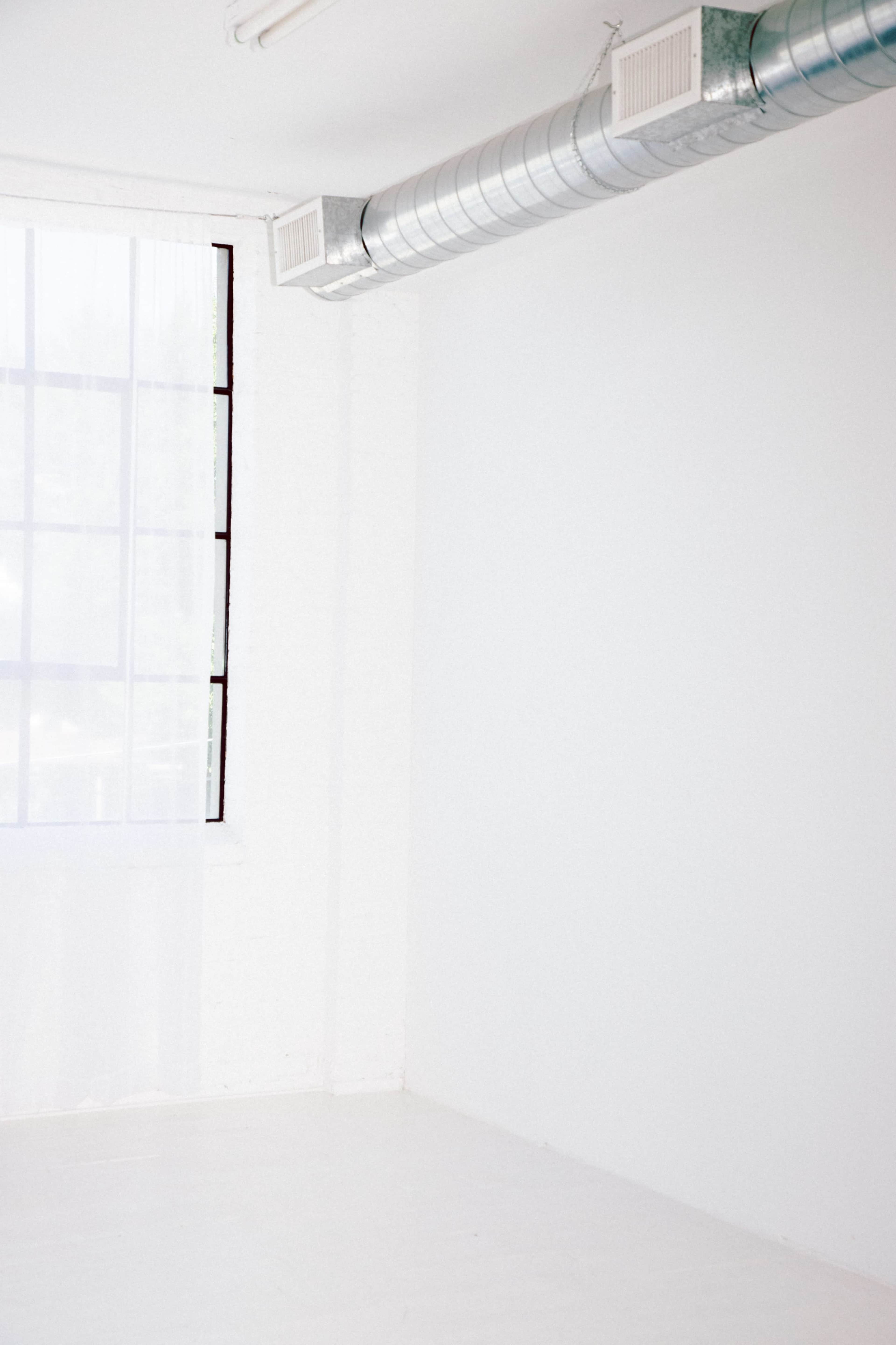 A bright, minimalist interior with a large window, exposed ductwork, and white walls and floors.