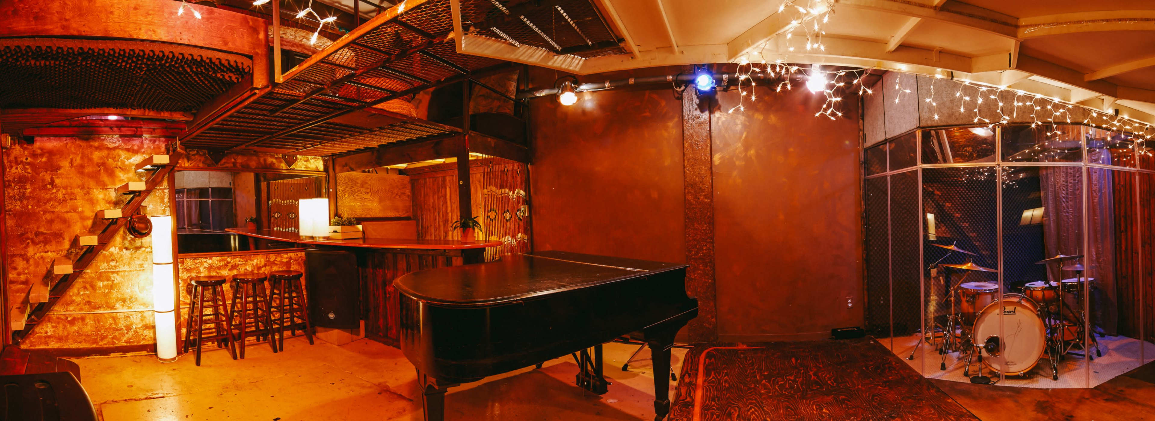 The image shows a dimly lit music venue with a grand piano, a bar with stools, and a section for a drum set, all framed by exposed brick walls and string lights.