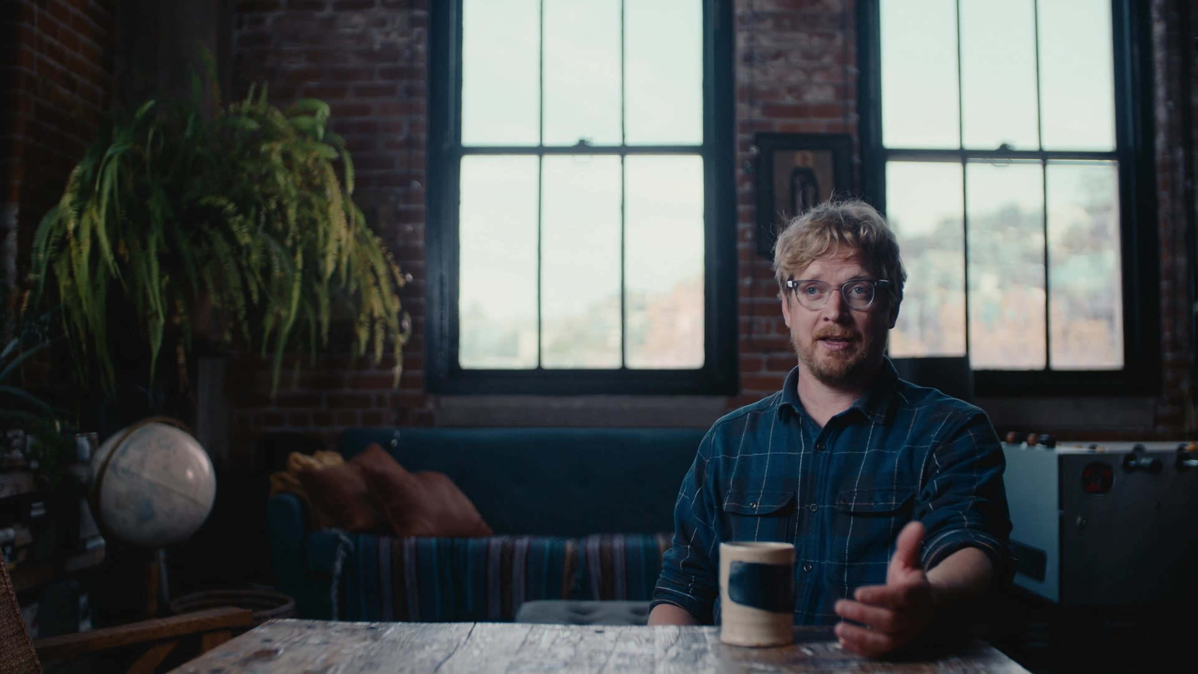A man with glasses sits at a wooden table in a room with large windows and a green plant, with a couch in the background.