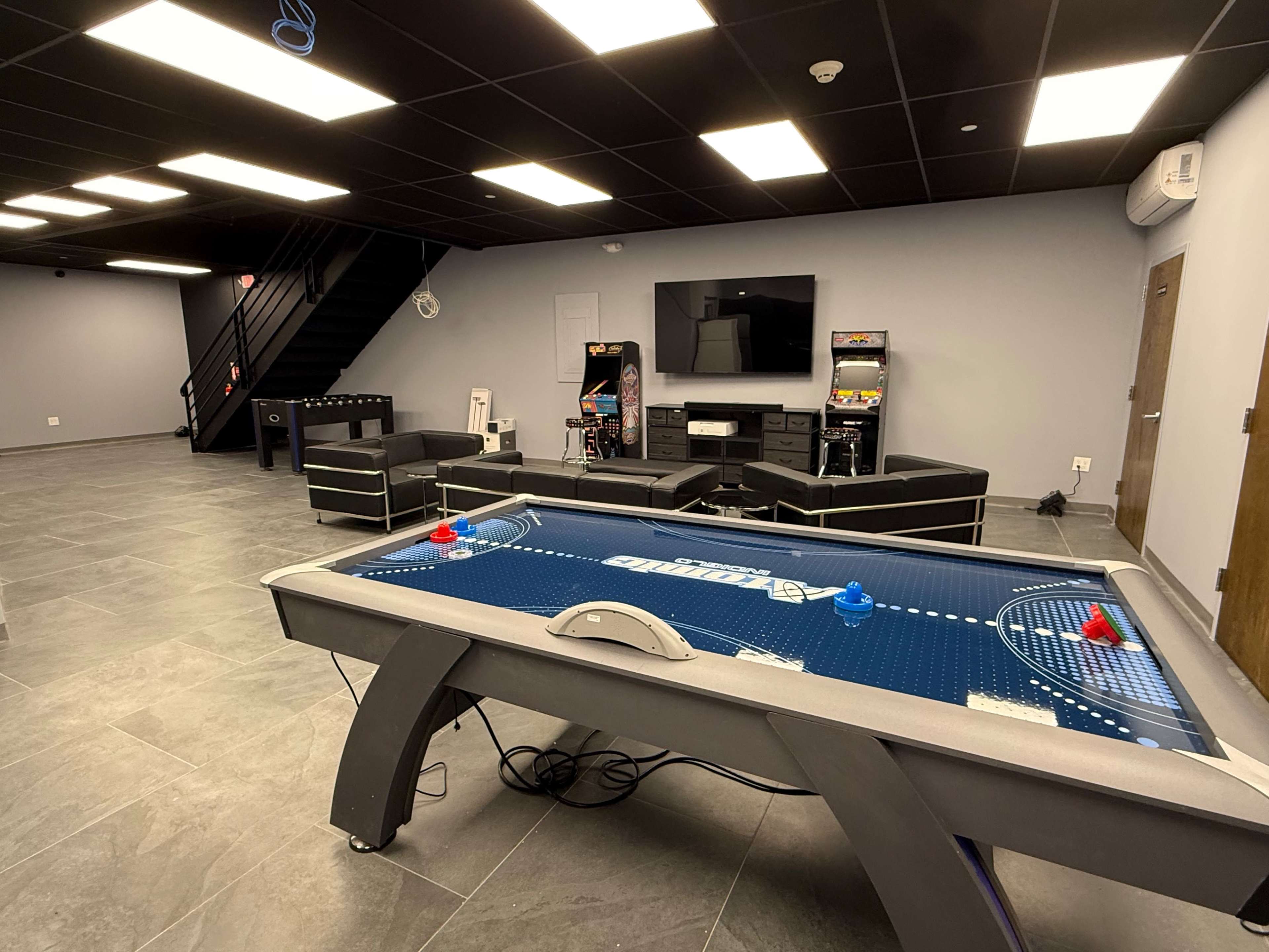 The image shows a game room featuring an air hockey table, a sitting area with black furniture, a television on the wall, and arcade machines in the background.