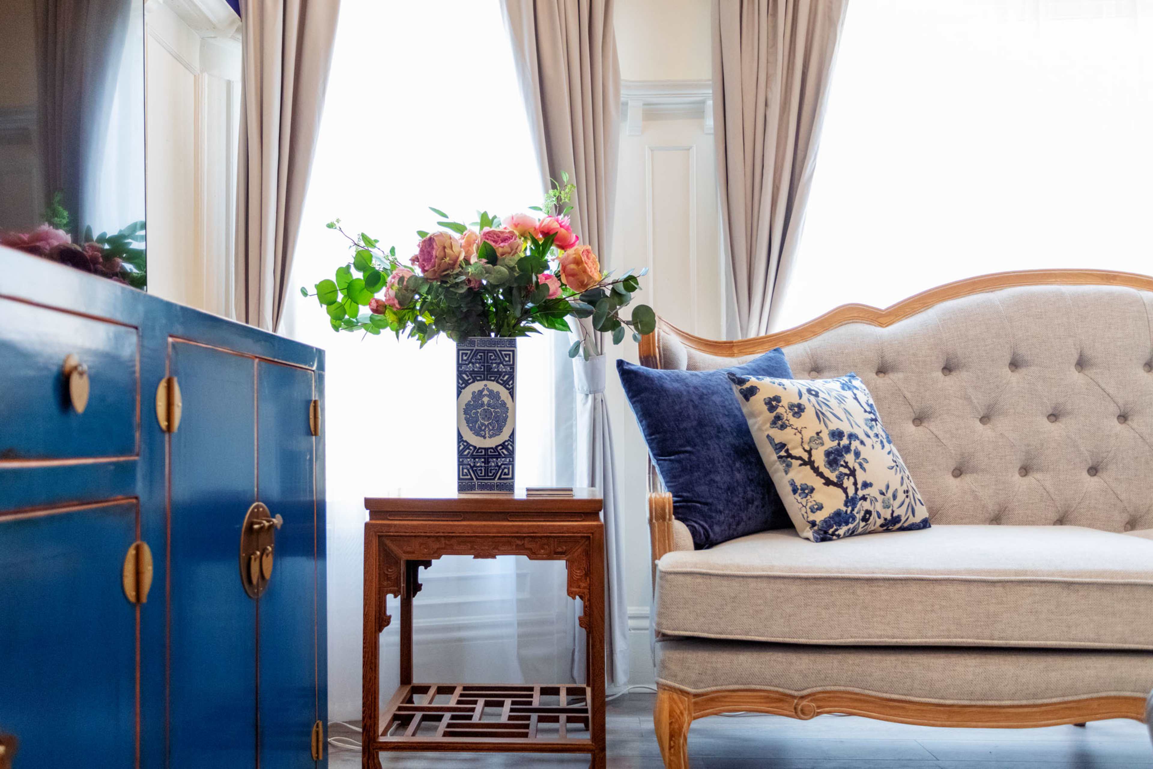A light-filled room features a blue cabinet, an ornate wooden side table, a gray tufted sofa with decorative pillows, and a vase of flowers on the table.