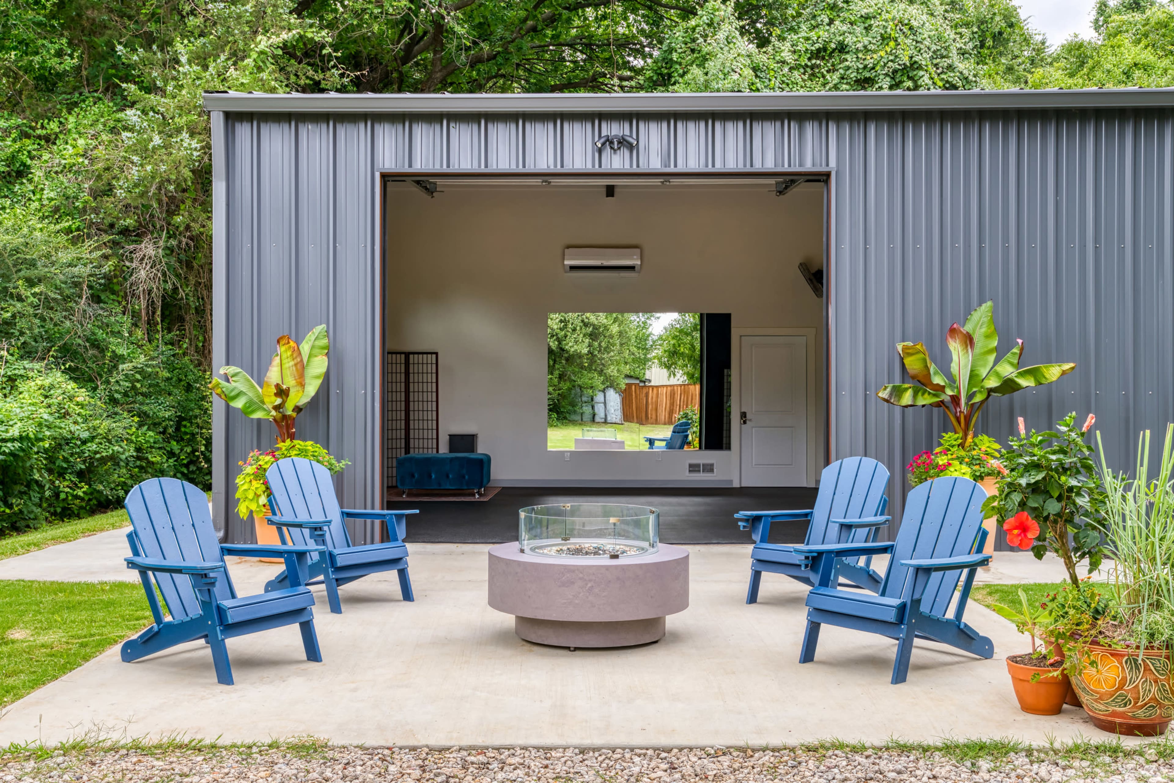 The image shows a modern outdoor seating area with four blue Adirondack chairs arranged around a circular fire pit, situated in front of a partially open garage with greenery in the background.