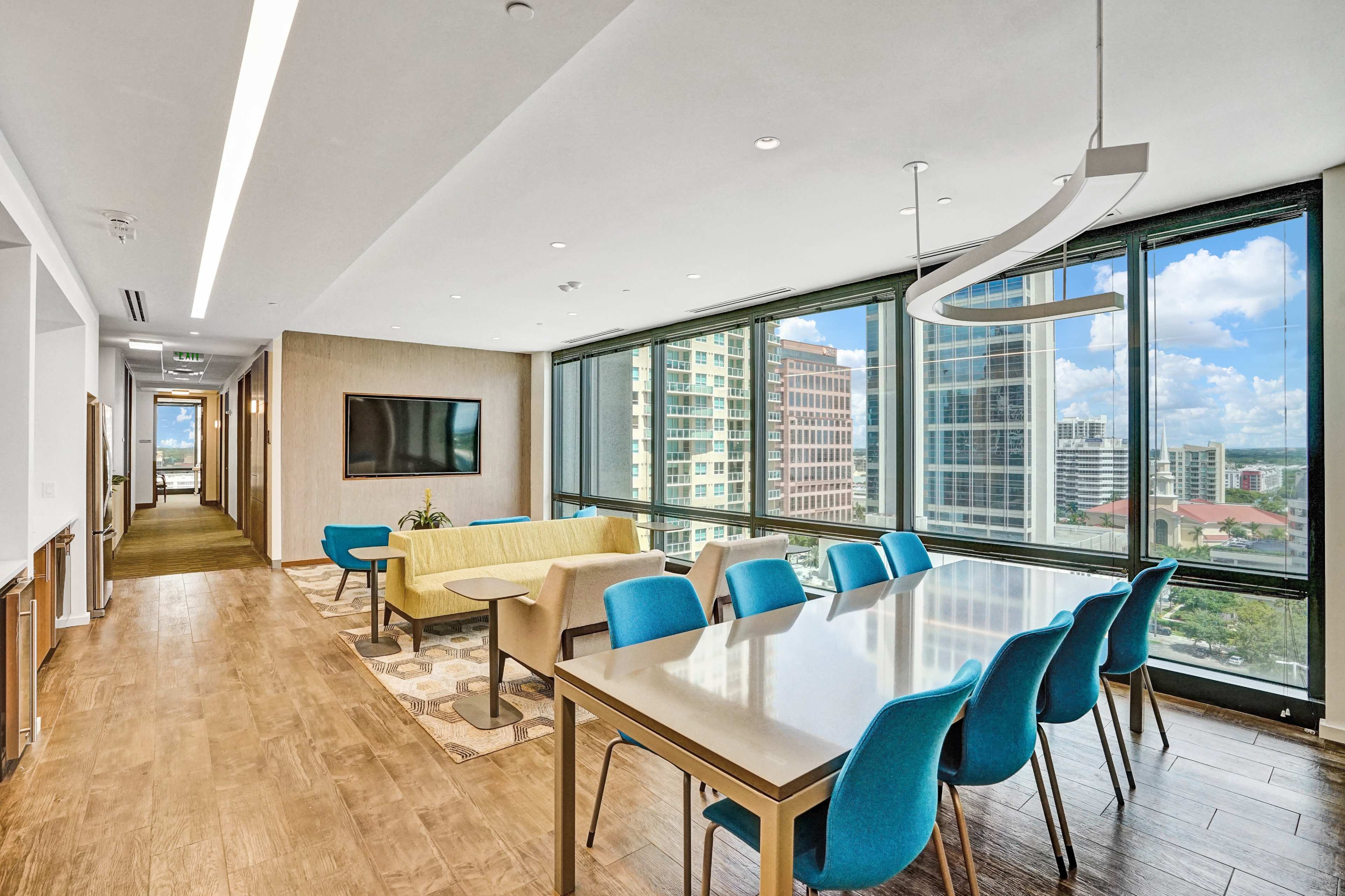 A spacious modern conference room features a long table with blue chairs, large windows offering city views, and a flat-screen TV mounted on the wall.