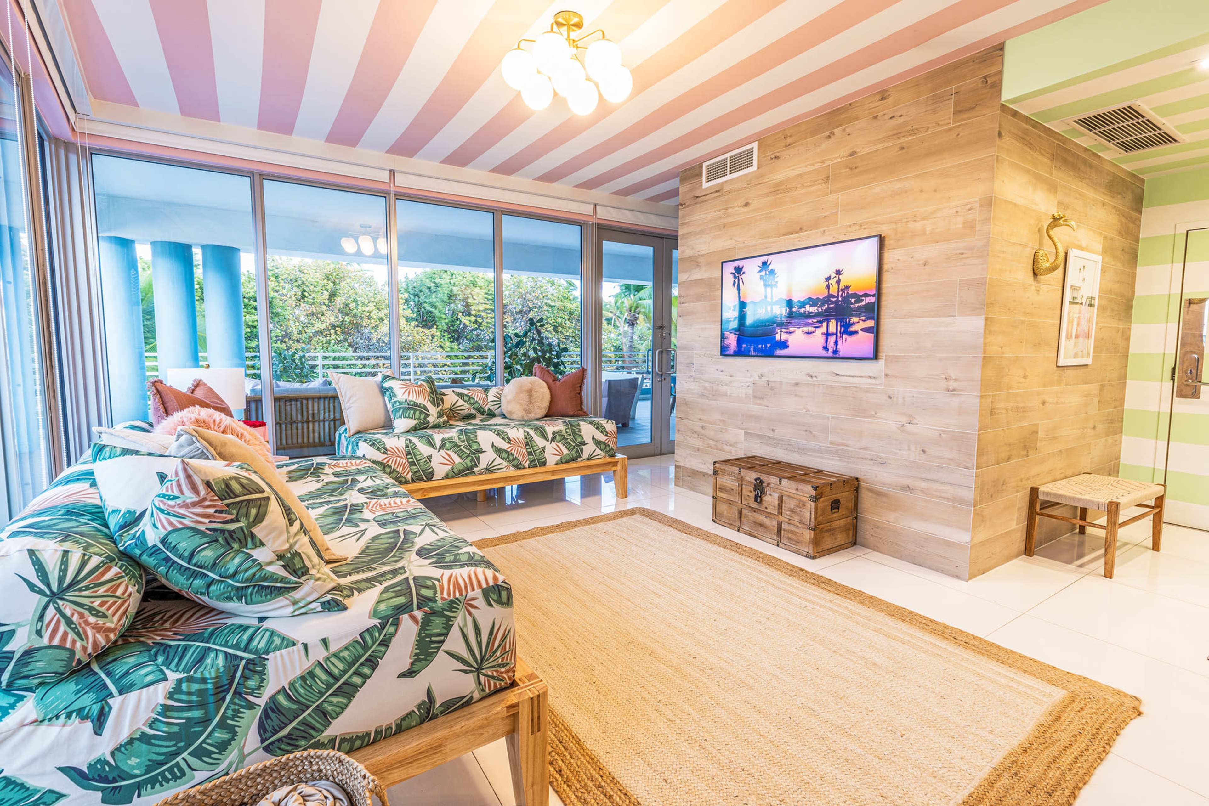 The room features two leafy-patterned daybeds, a wooden television stand, and large windows with views of greenery, all under a striped ceiling.