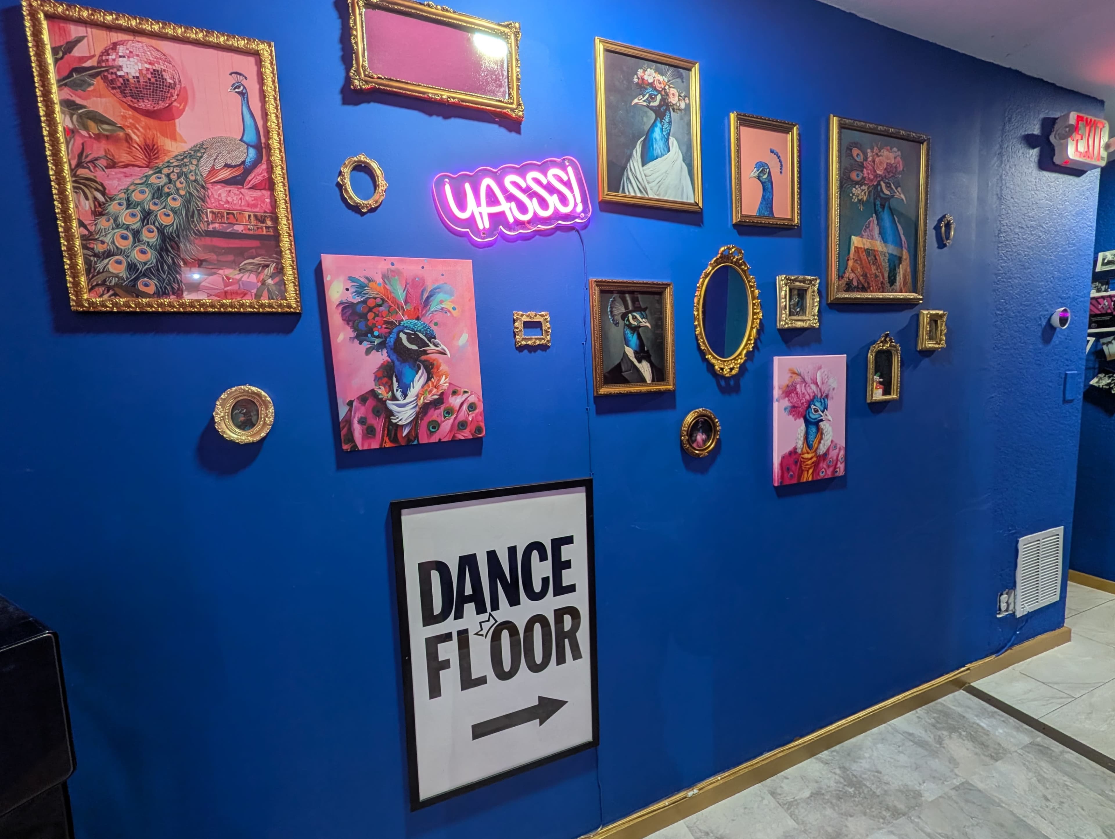 The image shows a vibrant blue wall adorned with various framed artwork featuring peacocks and decorative elements, along with a neon sign that reads "YASSSS" and a sign directing to the "DANCE FLOOR."