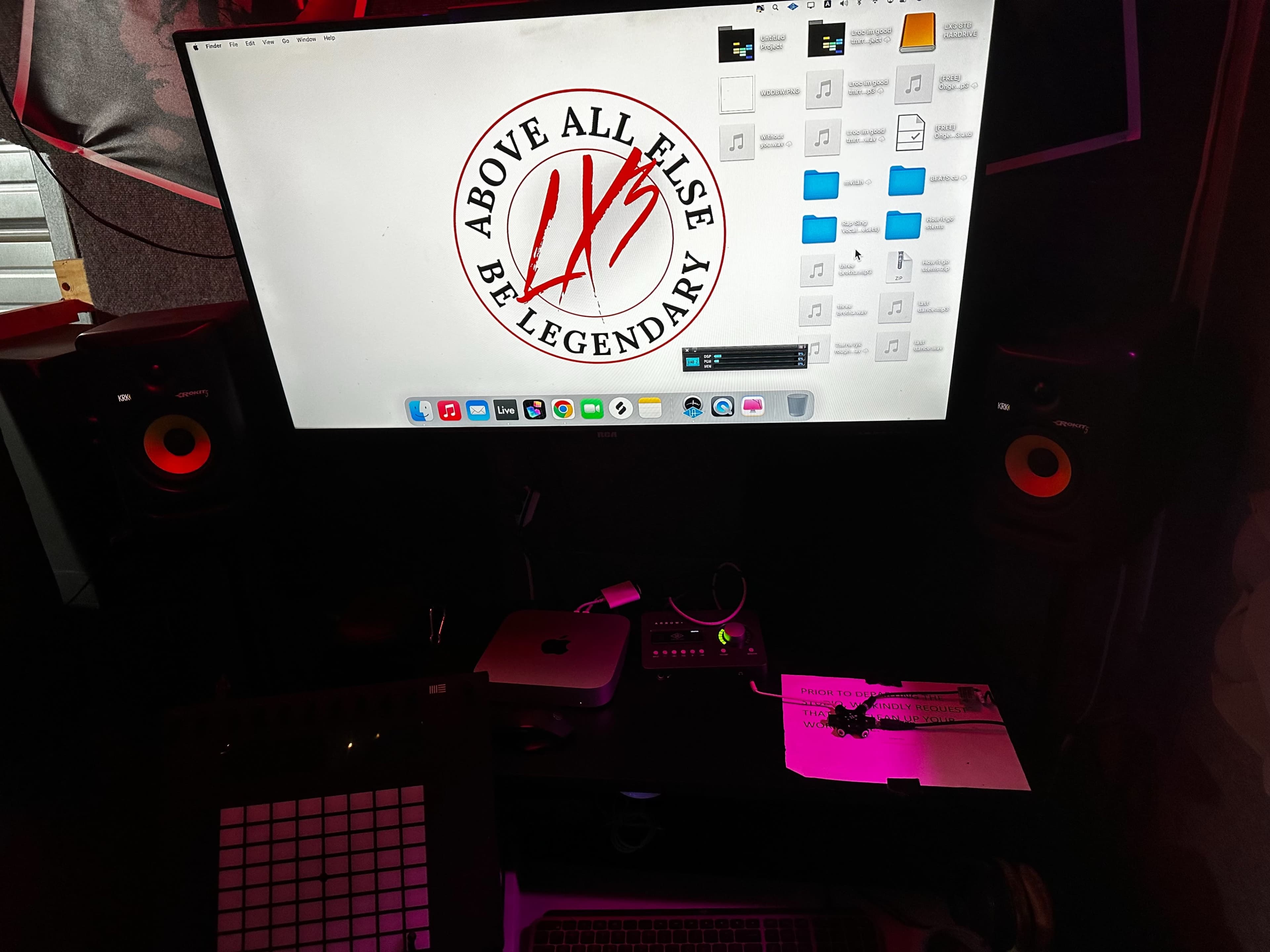 A computer monitor displaying a graphic with the phrase "Above All Else Be Legendary," positioned above a studio workspace featuring various audio equipment and a notepad.
