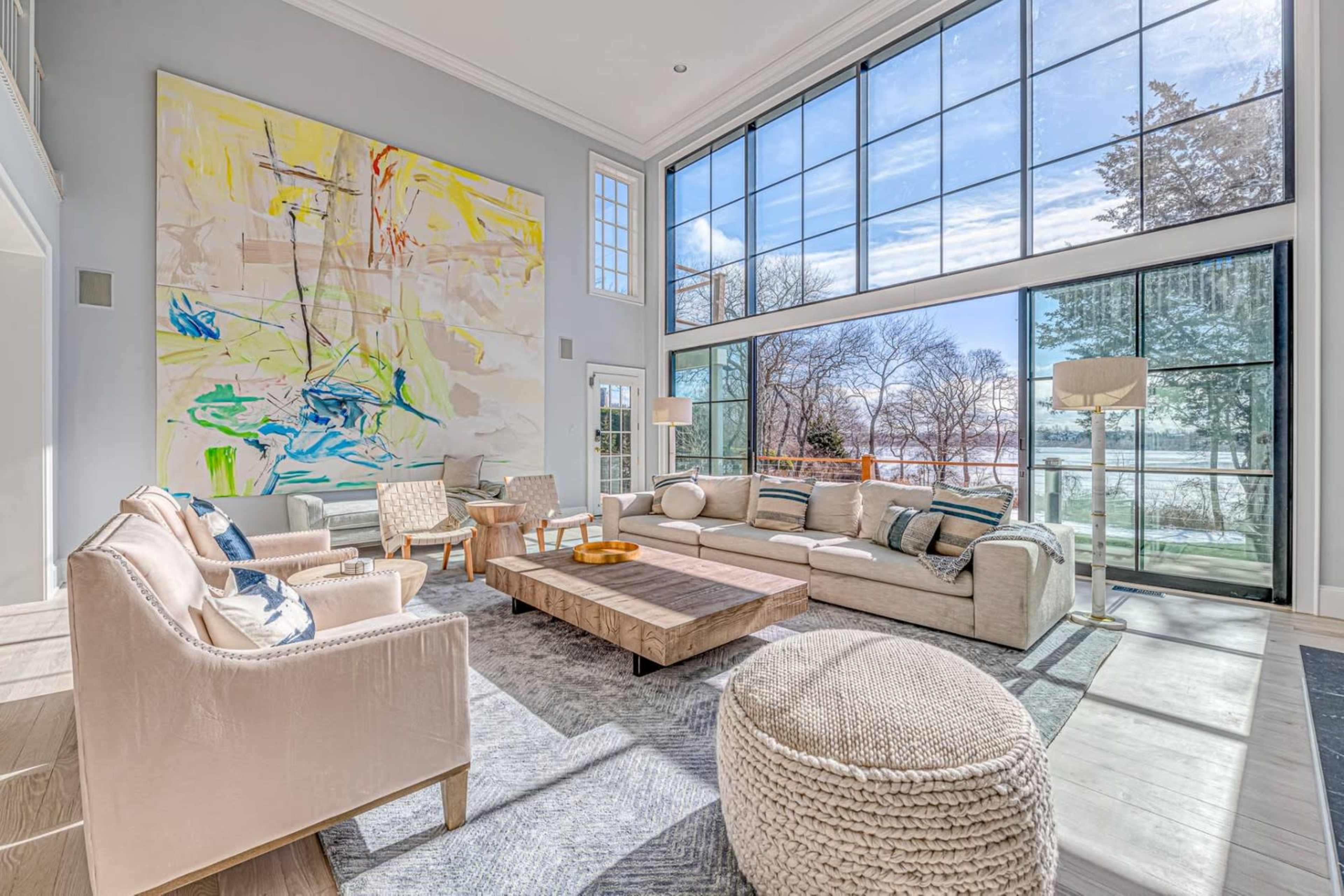 A bright living room features large windows with views of a lake, furnished with light-colored sofas, a wooden coffee table, and a prominent abstract painting on the wall.