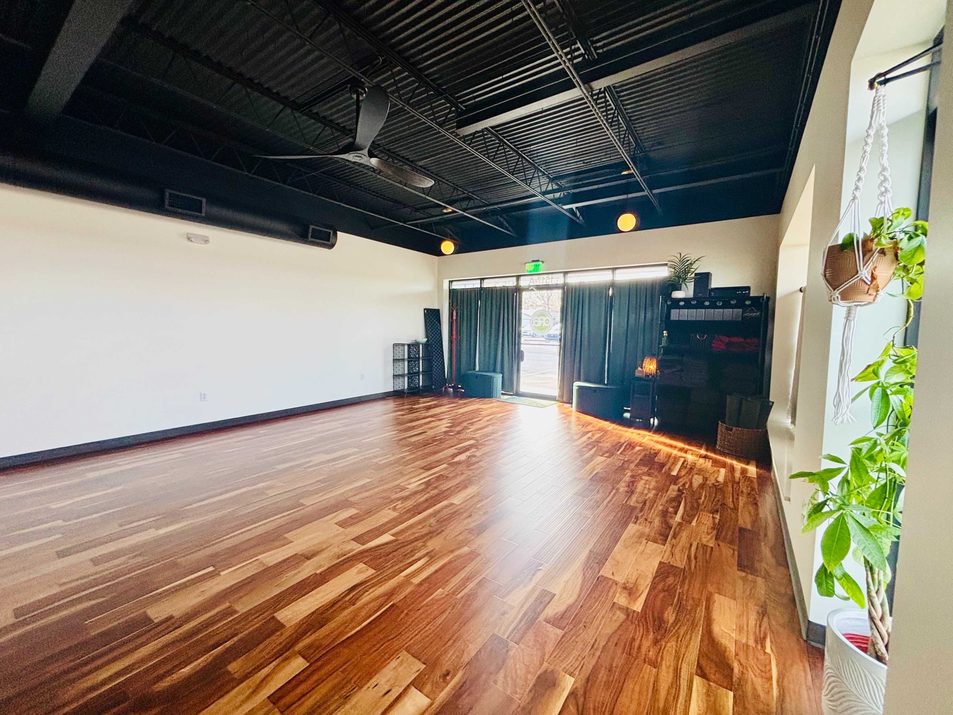 The image shows a spacious room with polished wooden floors, large windows, and minimal decor.