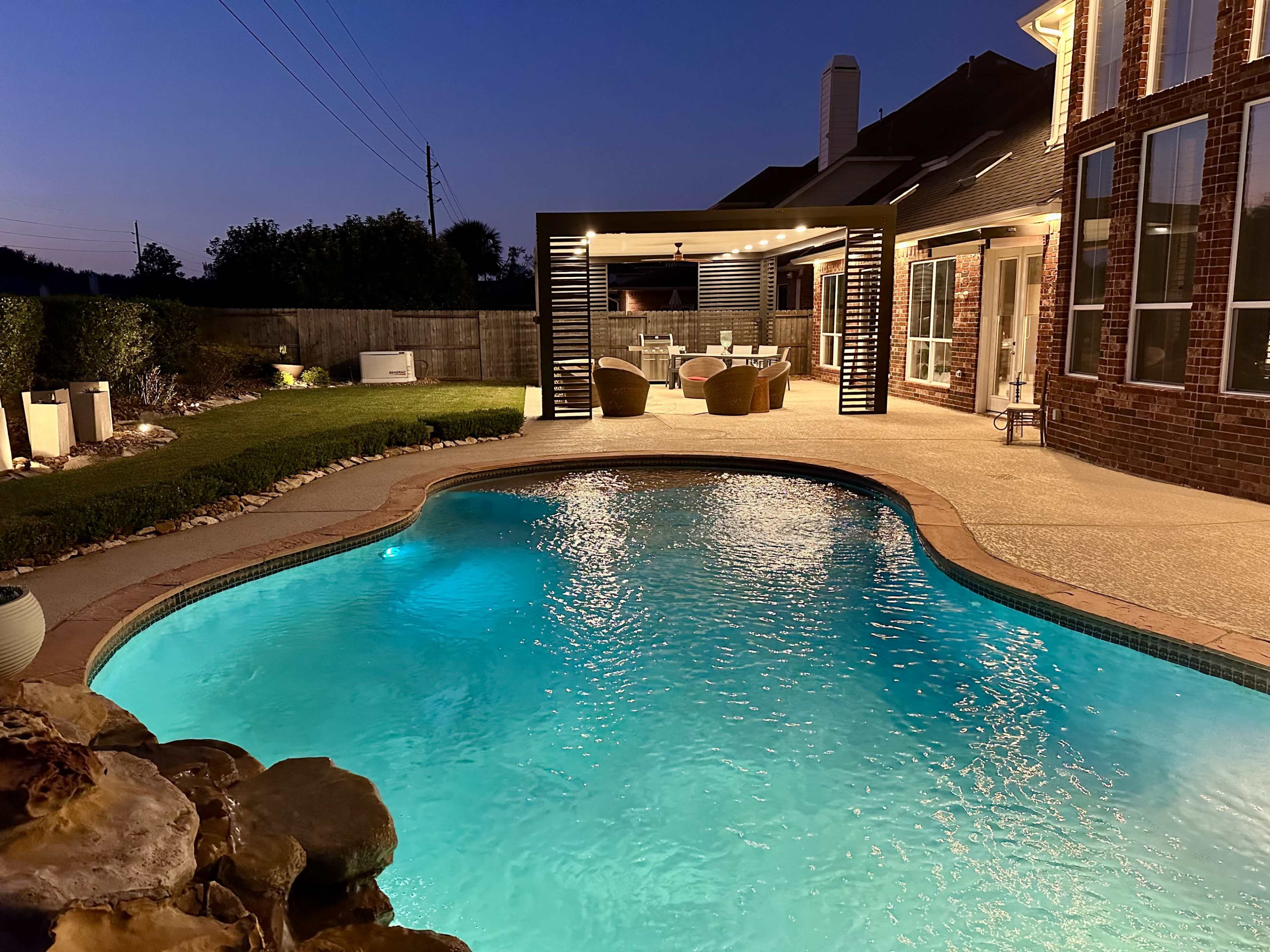 The image shows a backyard pool illuminated at dusk, with a covered outdoor seating area and well-maintained landscaping nearby.