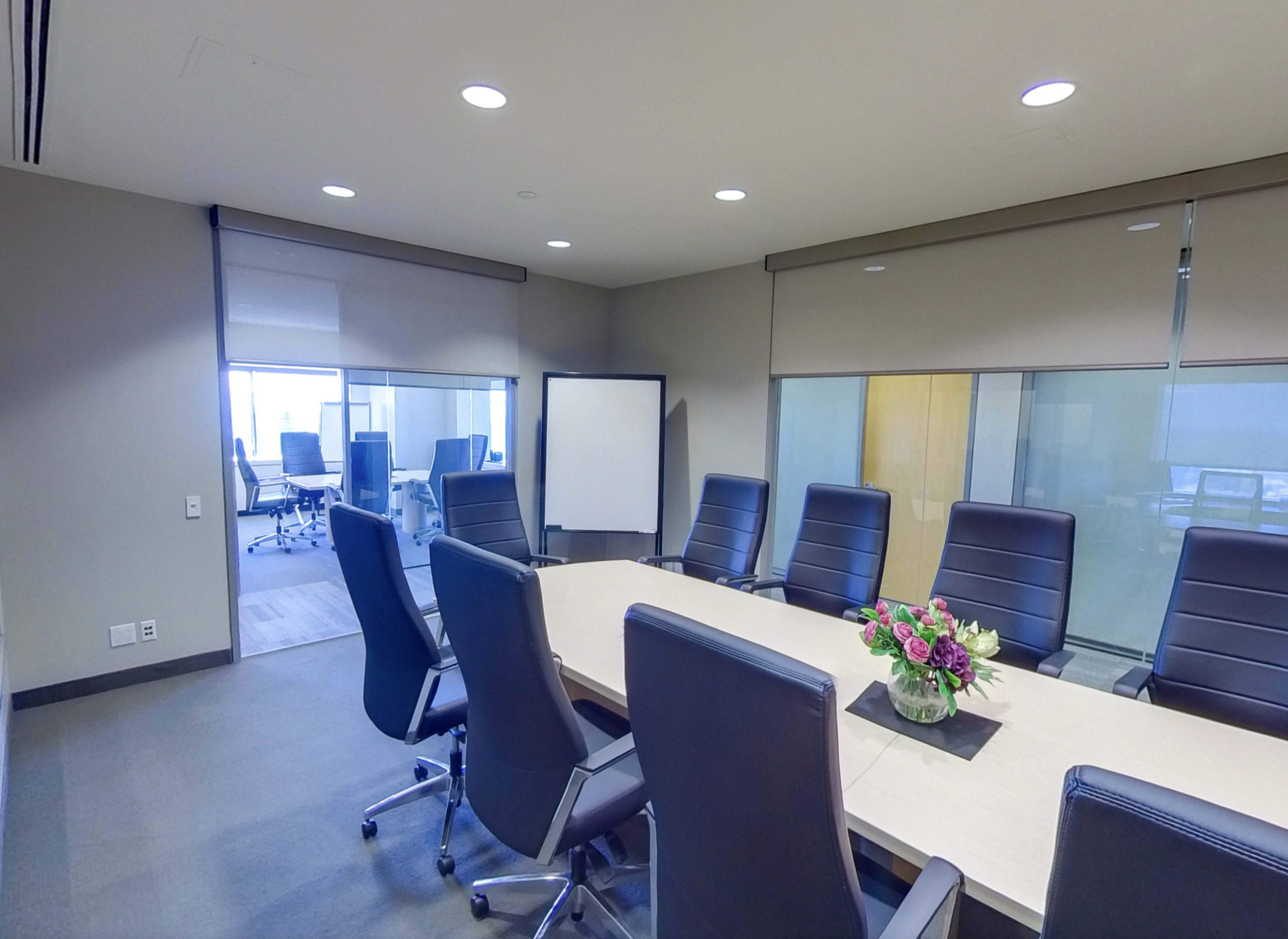 A modern conference room features a long table surrounded by black chairs, with a floral centerpiece and large windows showing additional meeting spaces.