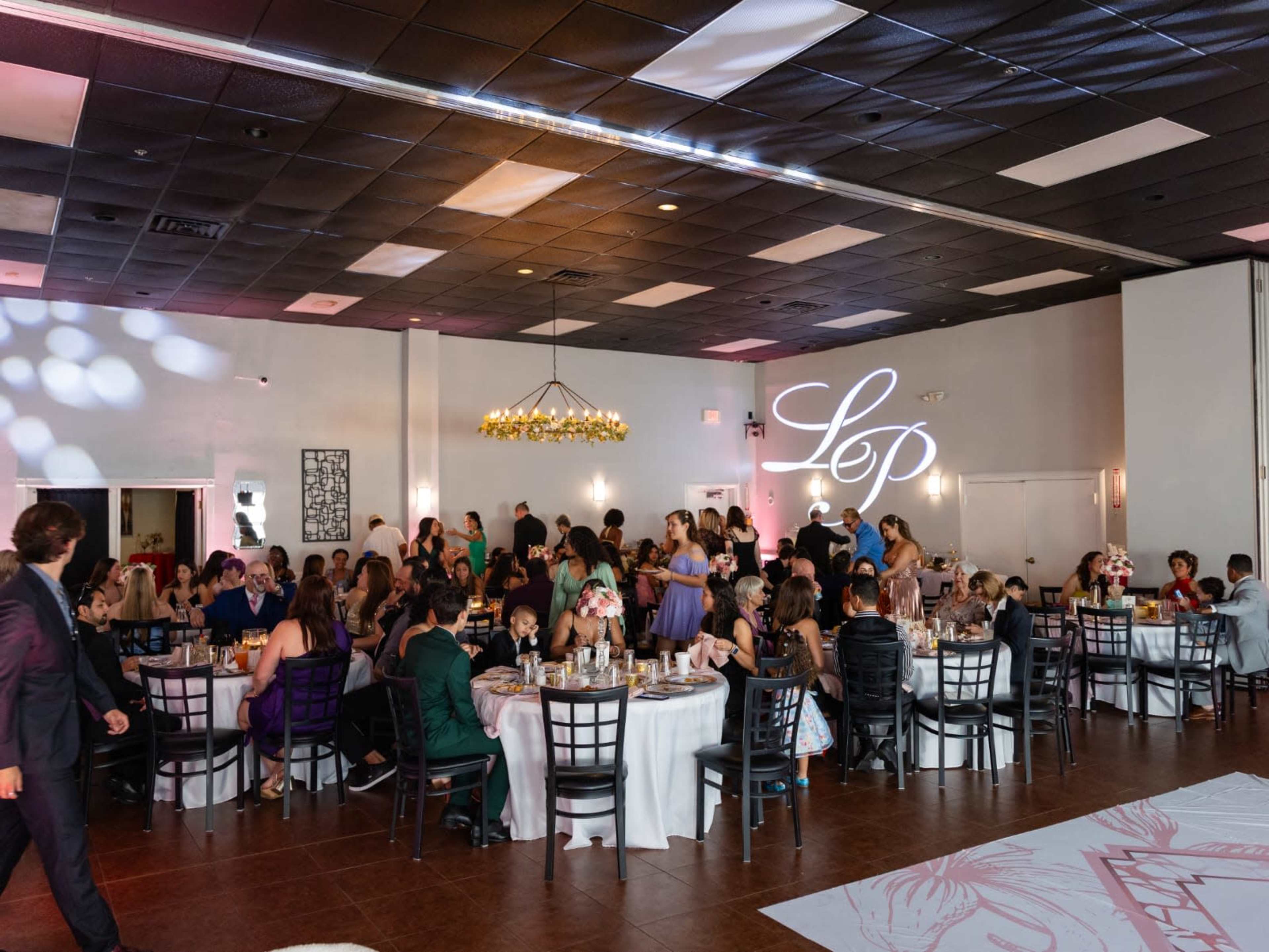 A large crowd of guests is gathered in a decorated banquet hall, seated at round tables for an event, while some people are mingling and a lighted monogram displays the name "Lep" on the wall.