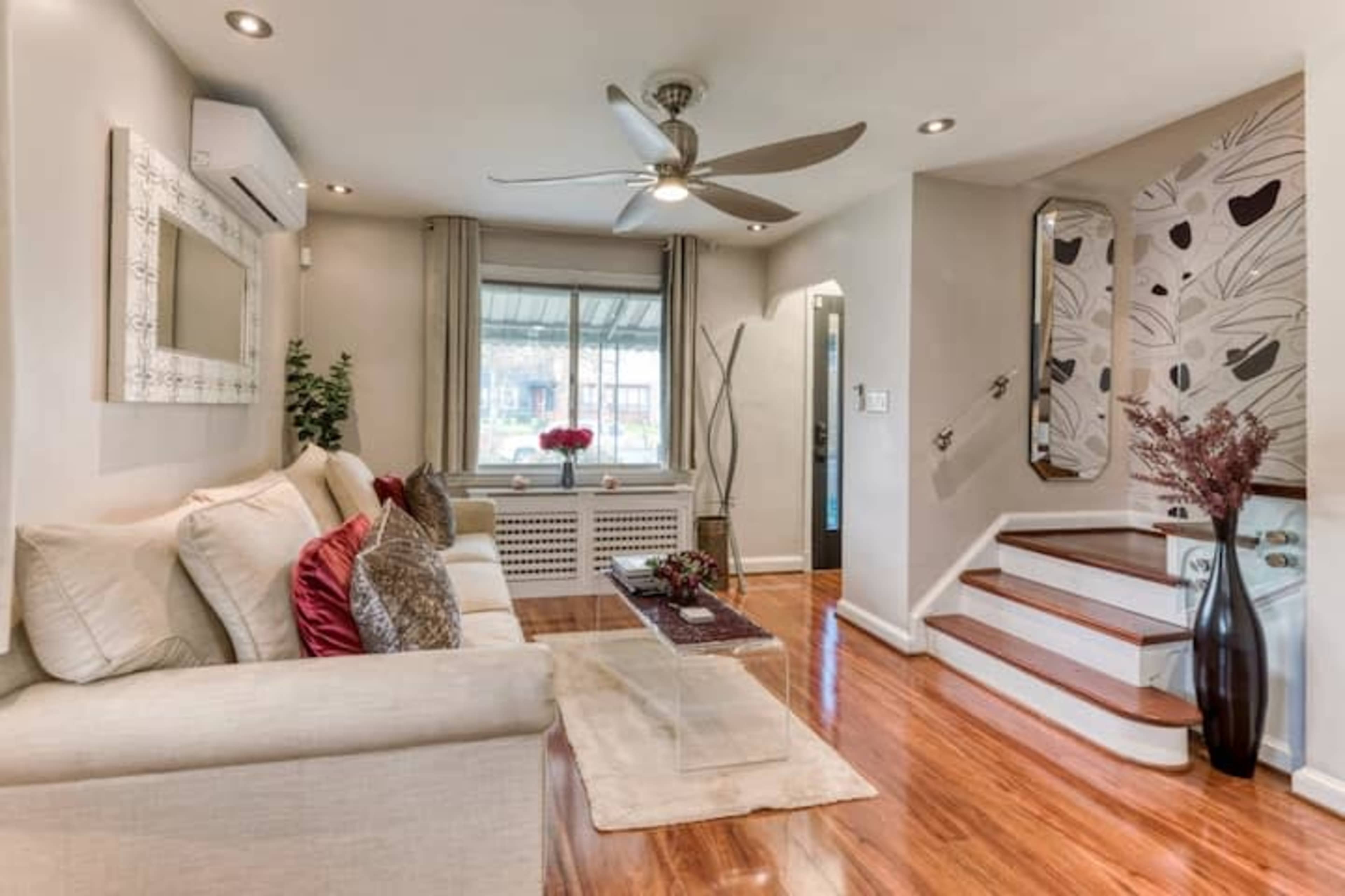 A bright living room features a cream-colored sofa with decorative pillows, a glass coffee table, and hardwood floors, alongside a staircase leading to an upper level.
