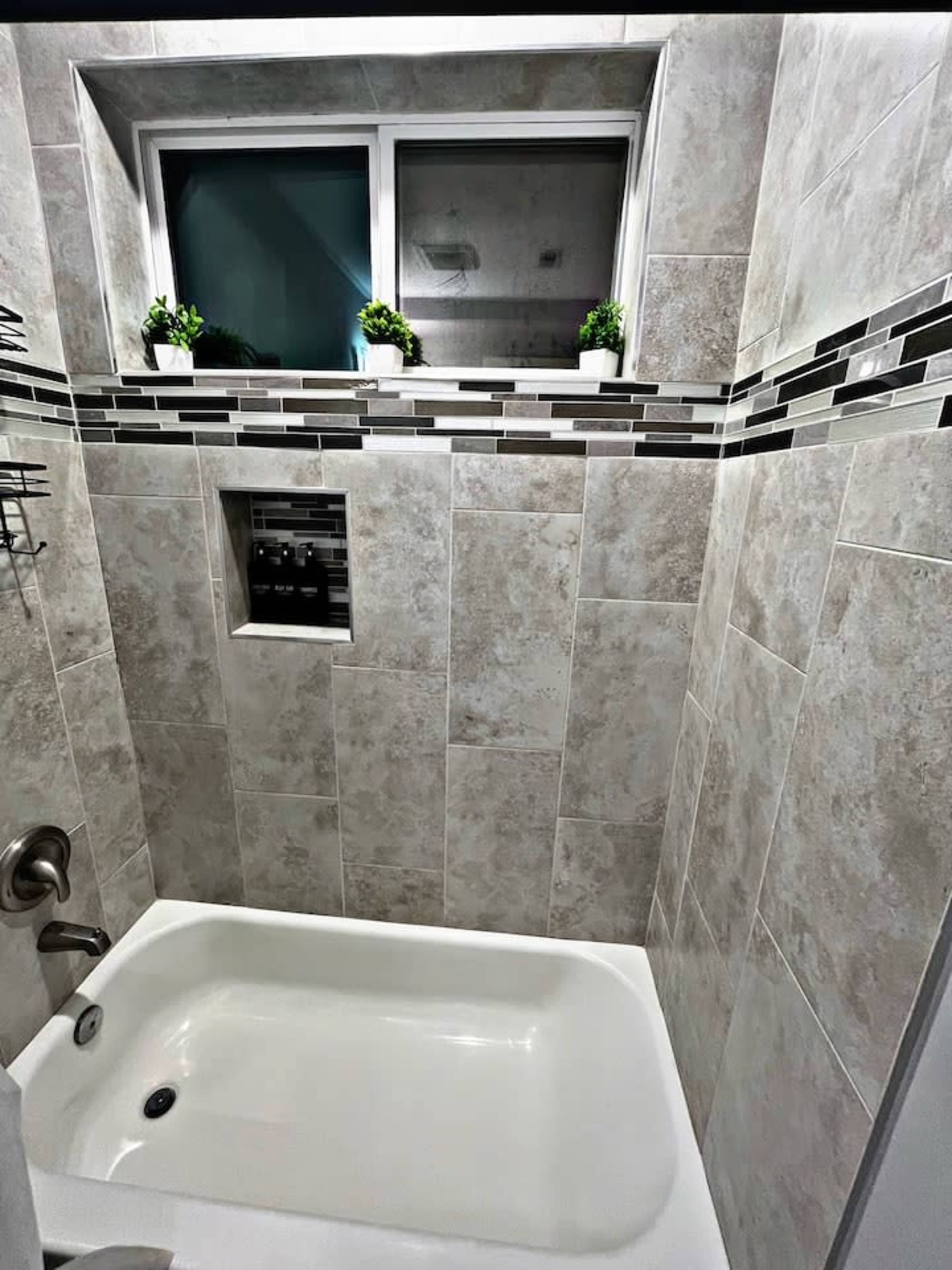 A modern bathroom with a bathtub, tiled walls, and a window above the tub featuring small decorative plants.