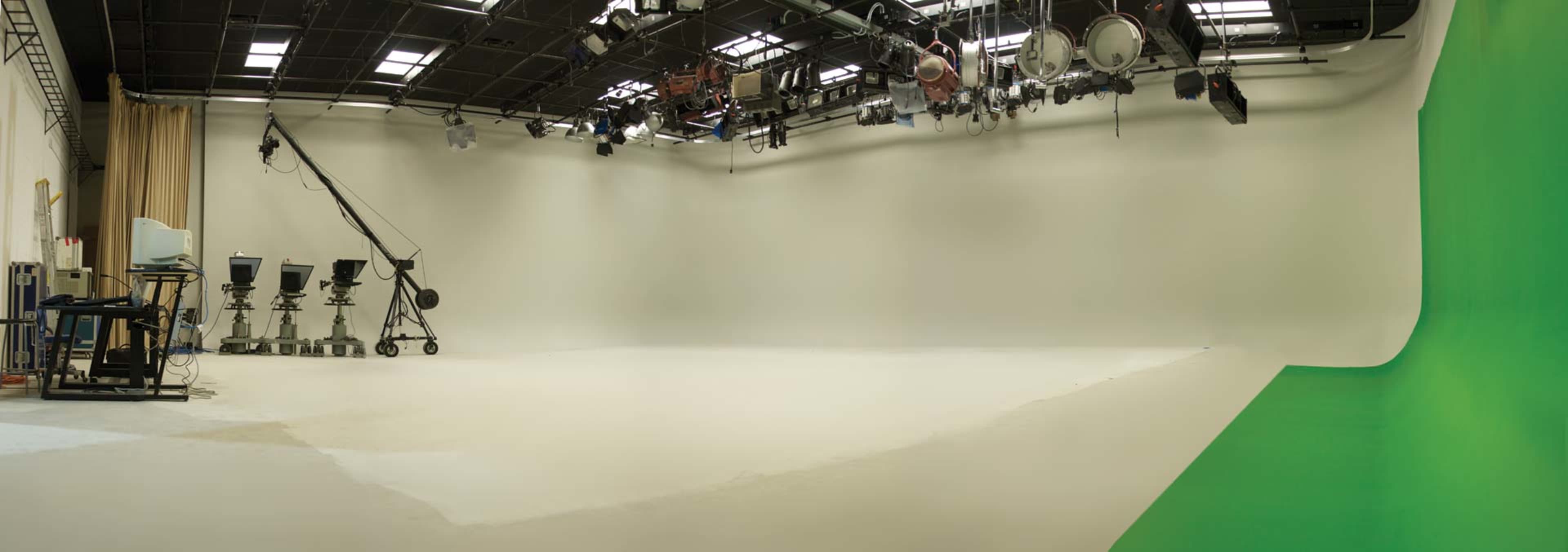 A spacious film studio with a white backdrop and a green screen on one side, equipped with lighting and camera equipment.