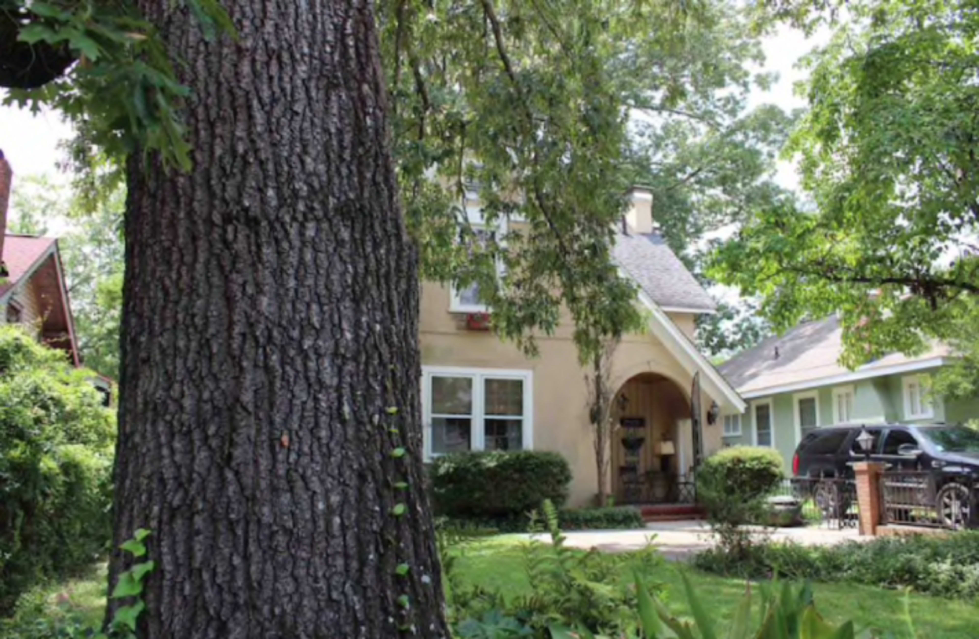 A two-story house with a light-colored facade and a sloped roof is situated behind a large tree in a residential area.