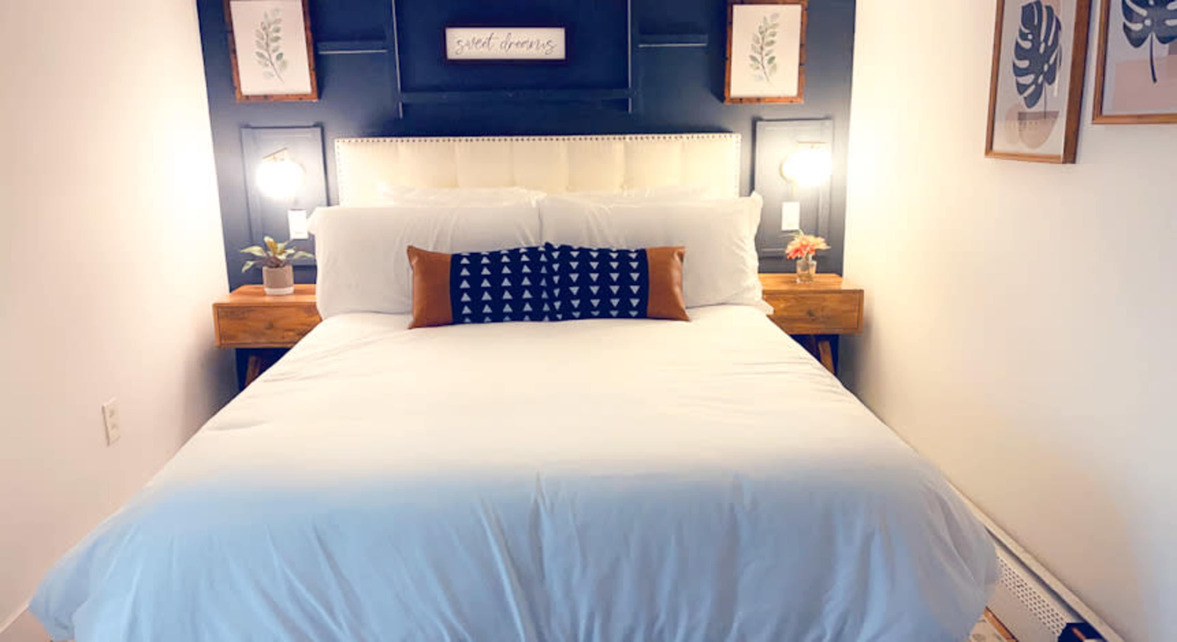 A neatly arranged bedroom with a white bedspread, decorated with a dark blue accent pillow, flanked by wooden nightstands and framed botanical prints on the wall.