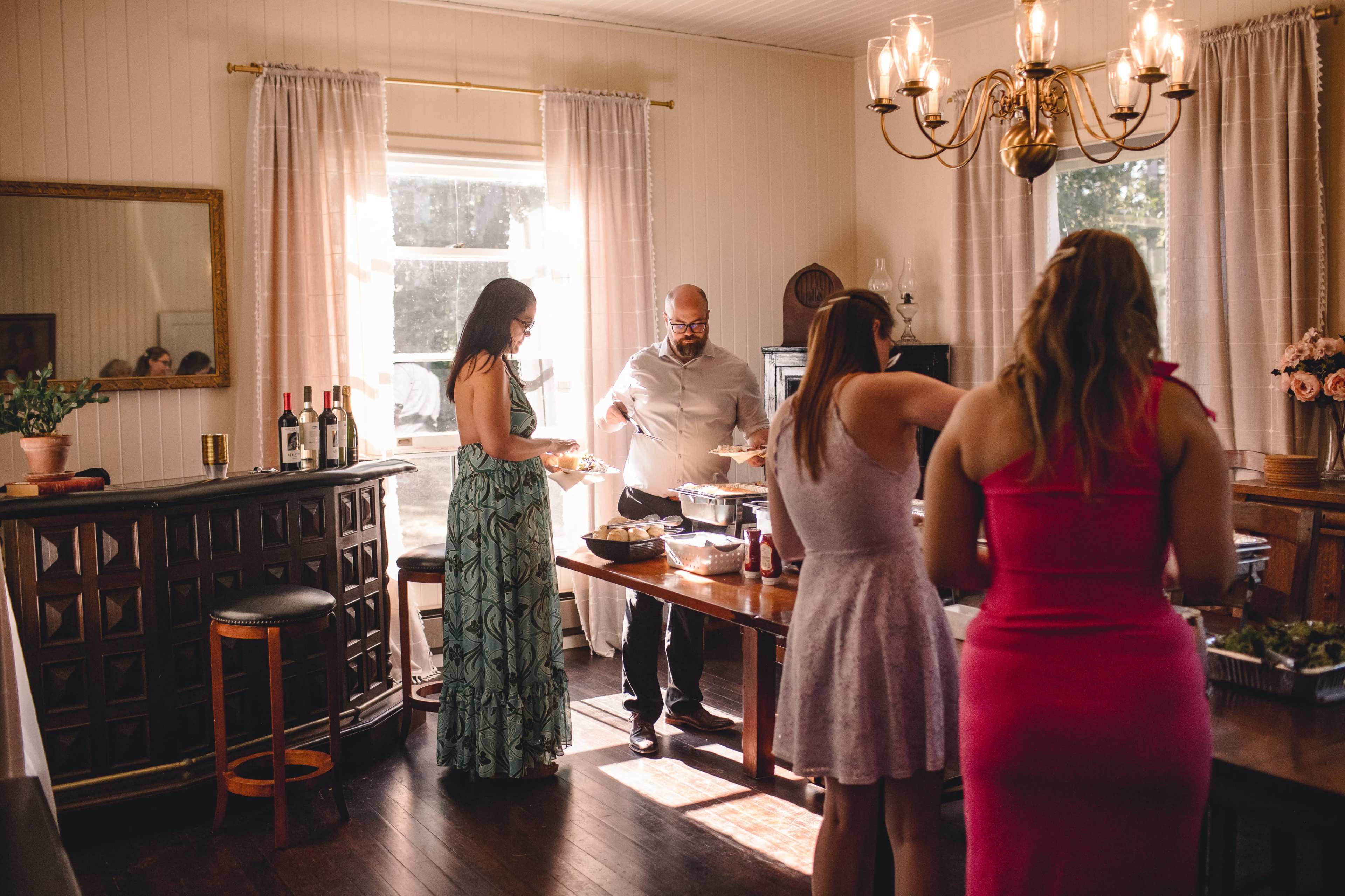 A group of people is gathered in a well-lit dining room, serving and preparing food from a table while a bar is visible in the background.