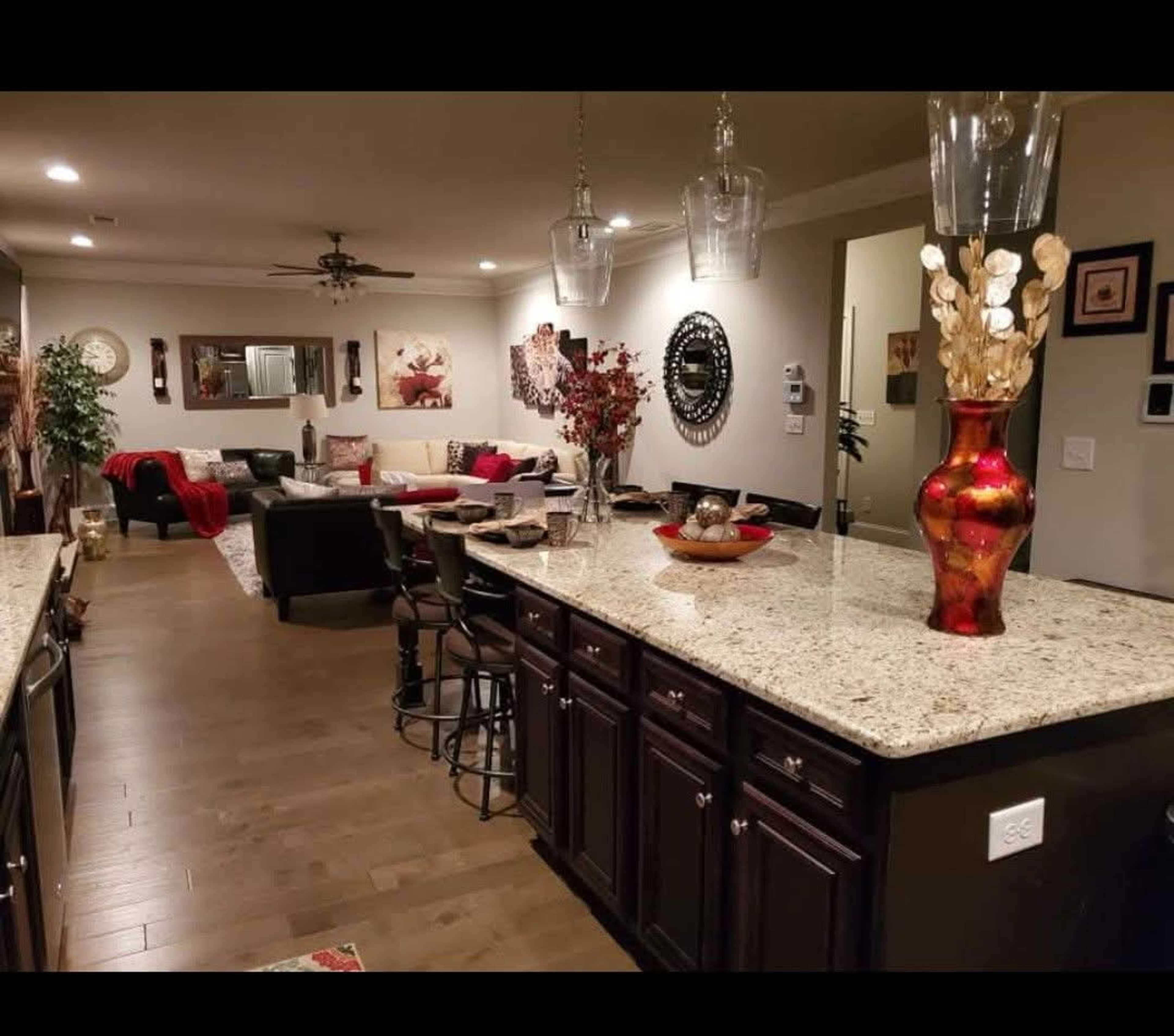 The image shows a spacious kitchen leading into a living area, featuring dark wooden cabinets, a granite countertop, and decorative elements such as a red vase and plants.