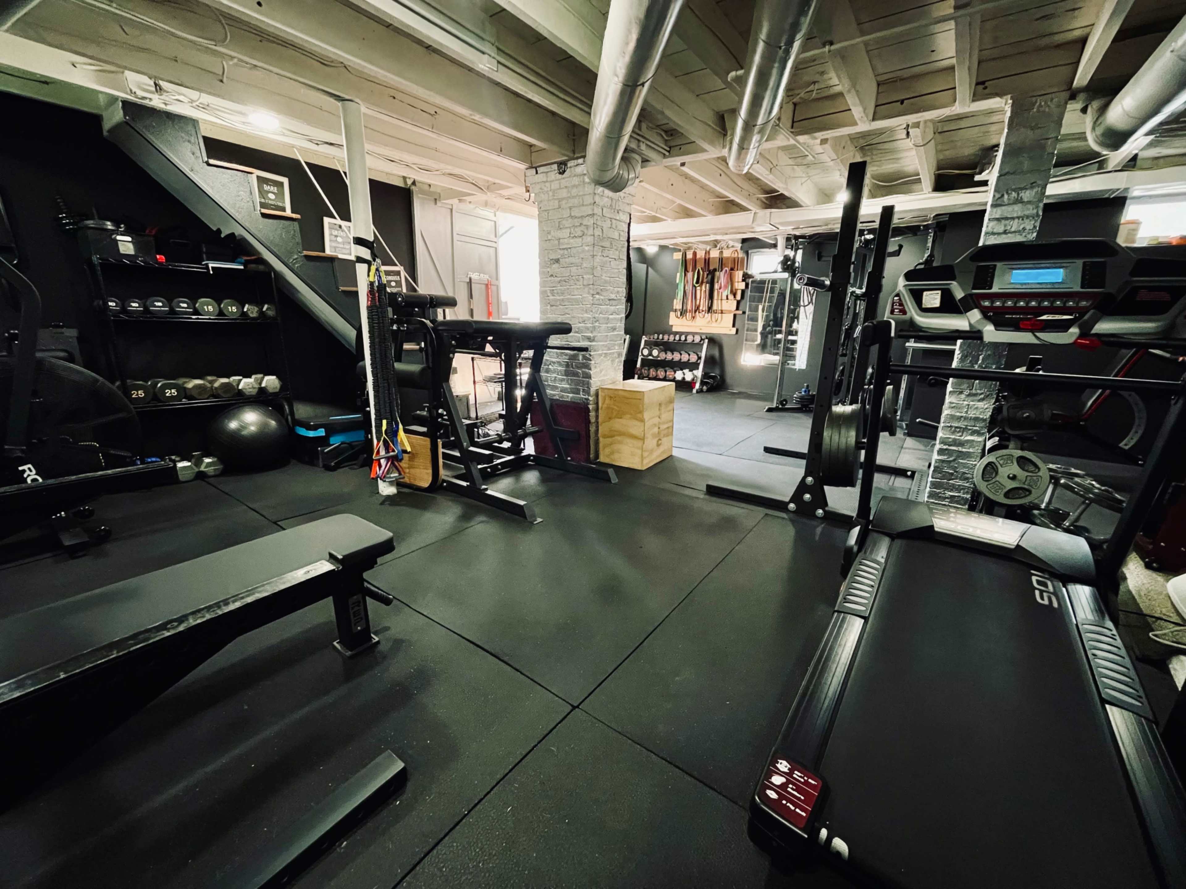 A home gym with various exercise equipment, including dumbbells, a treadmill, and weightlifting benches, set on black rubber flooring.