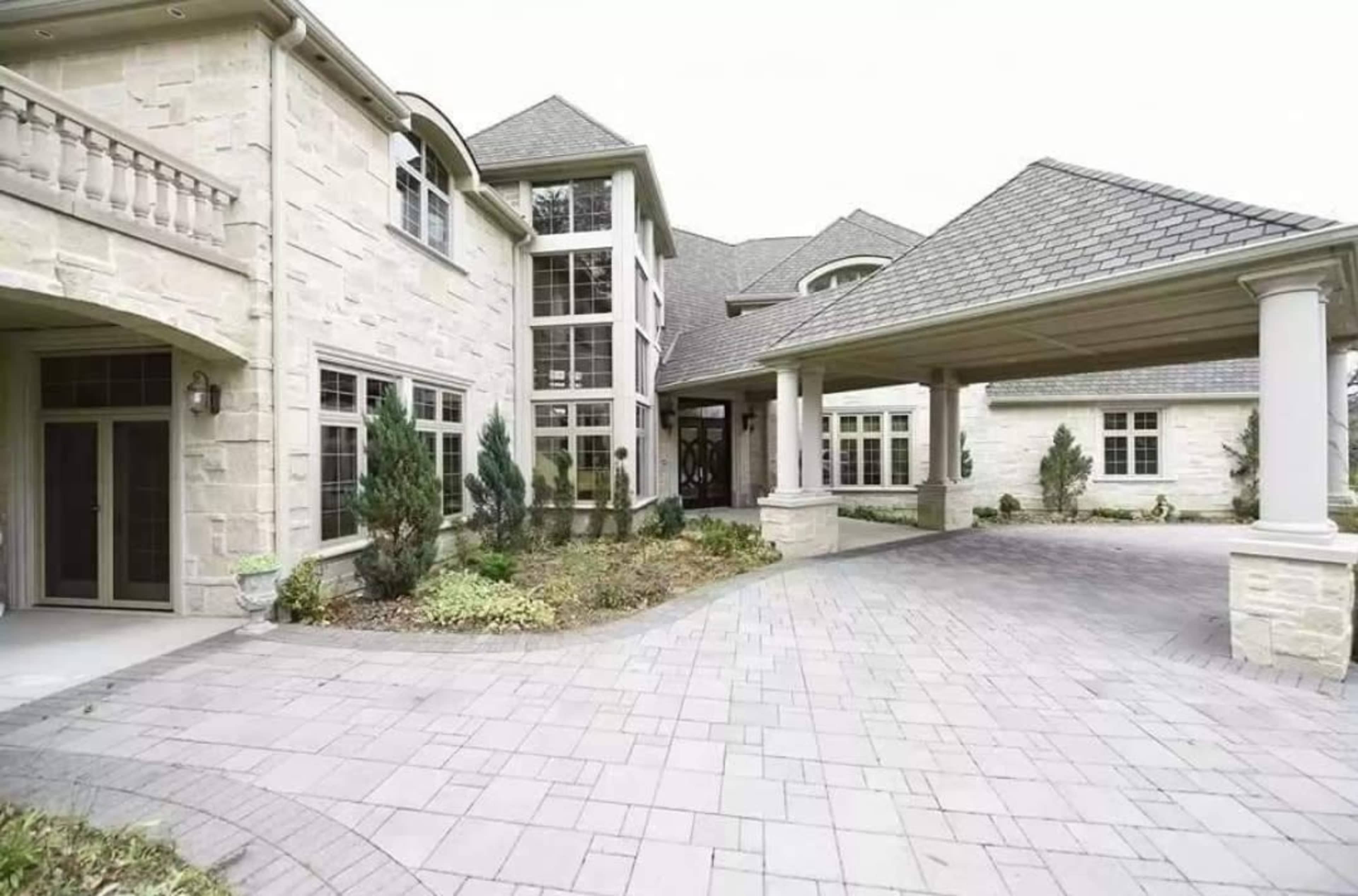 A large stone house with landscaped gardens and a circular driveway, featuring multiple windows and a covered entrance.