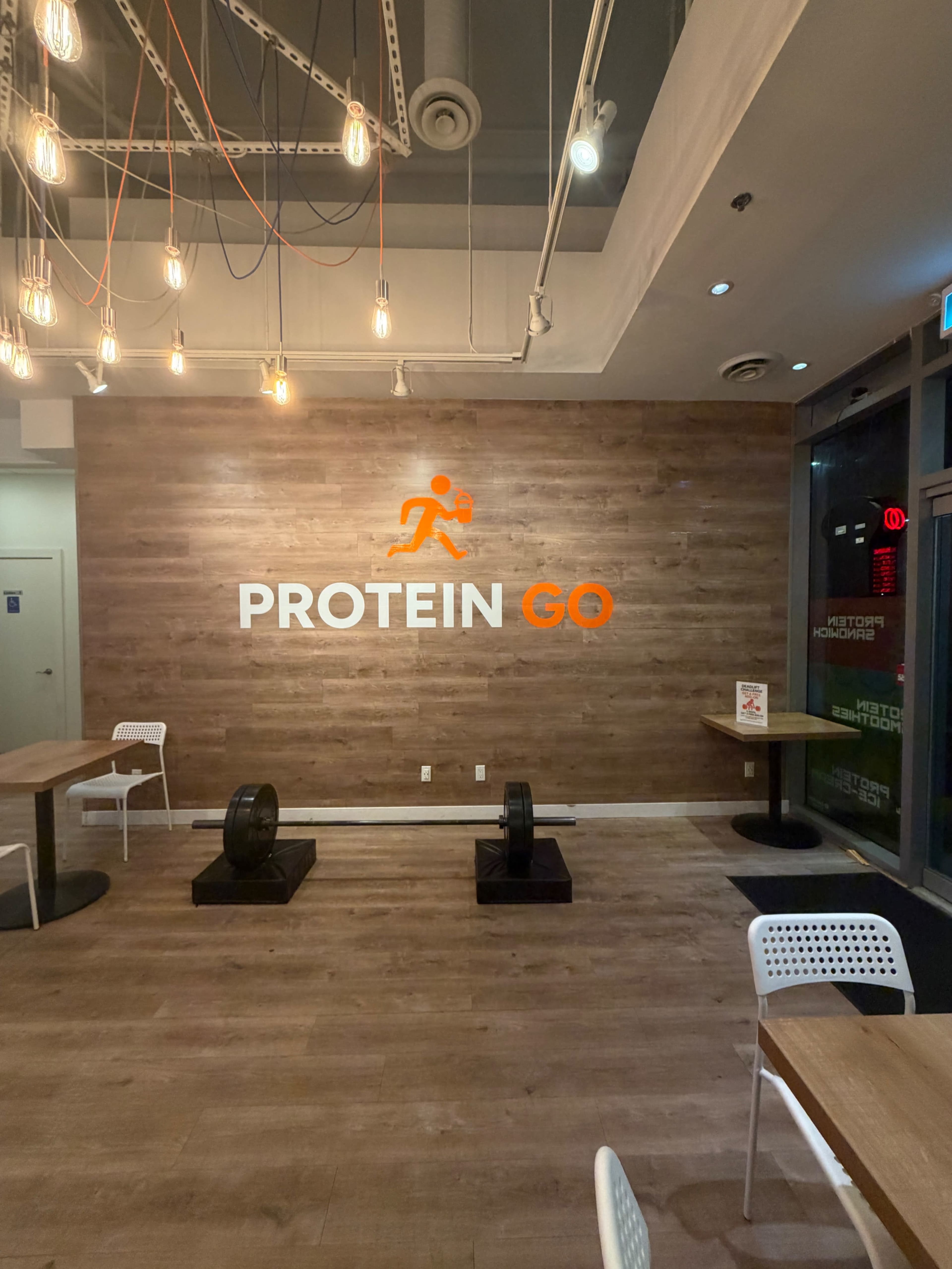 The image shows an interior of a fitness-themed establishment with a wooden wall featuring the logo "PROTEIN GO" and a couple of weightlifting plates on the floor.