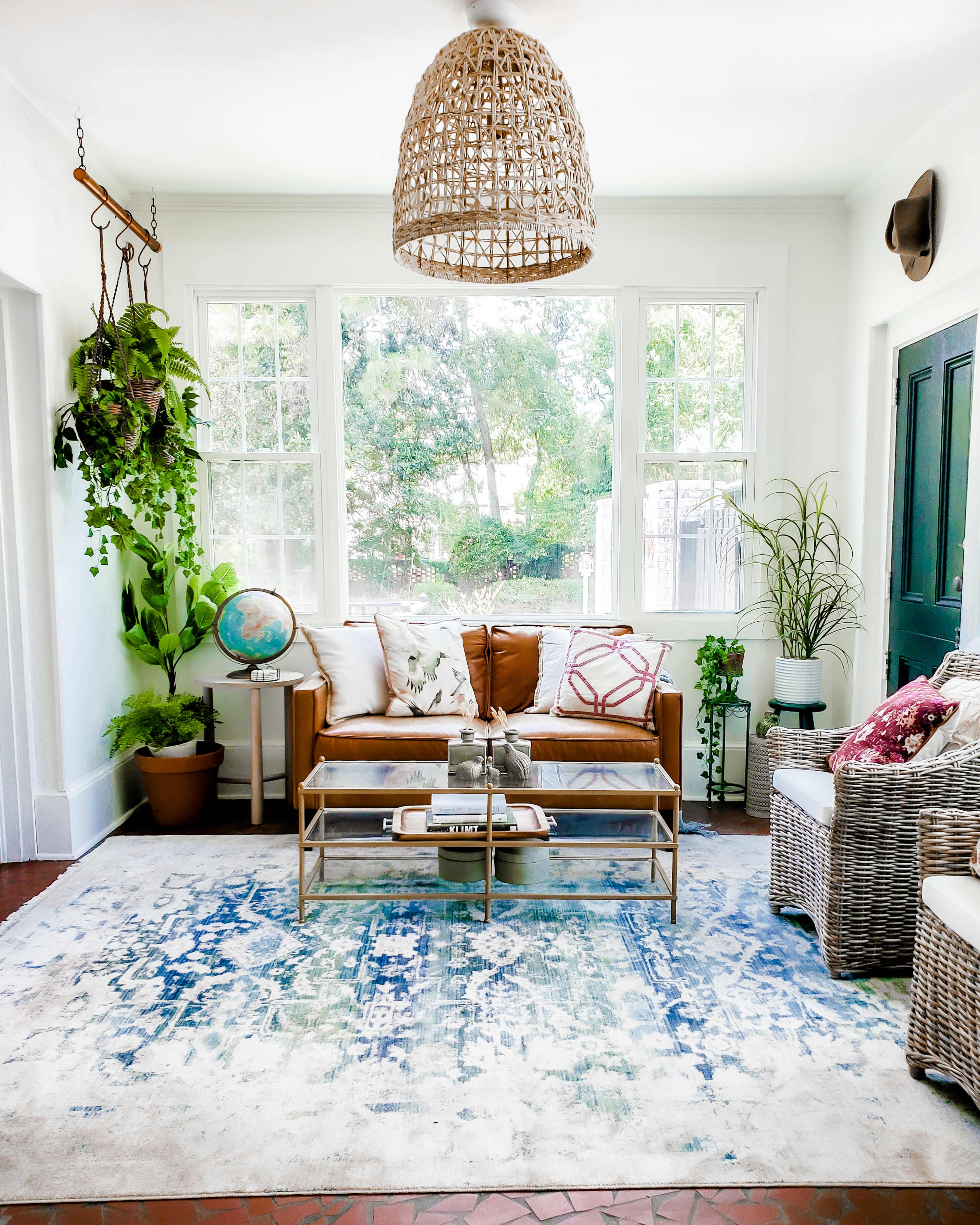 A bright living room features a brown leather sofa, two white armchairs, a coffee table, and decorative plants, all set against large windows that overlook greenery outside.