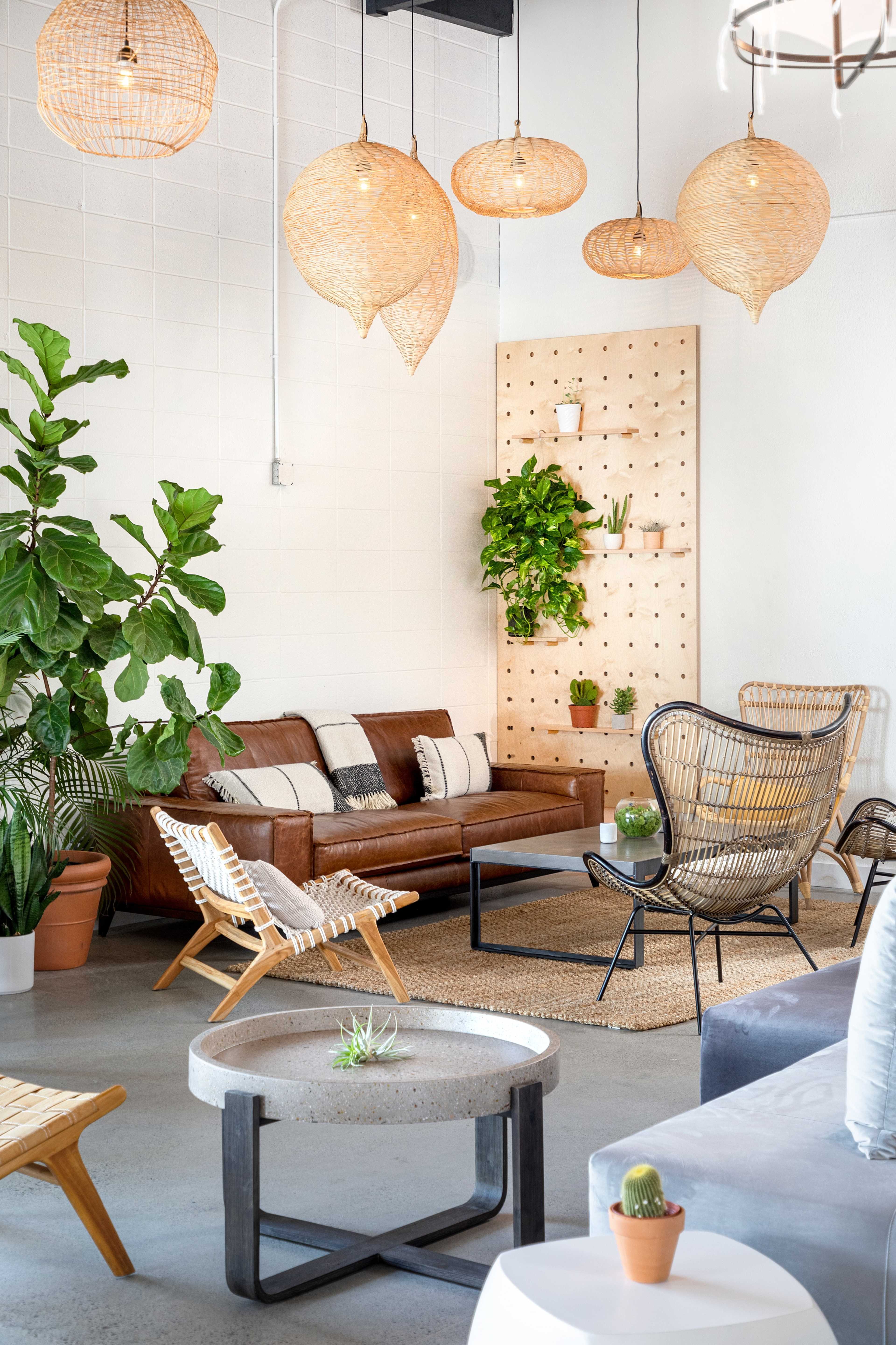 A modern, airy living space featuring a brown leather sofa, woven pendant lights, and various plants arranged around a coffee table.