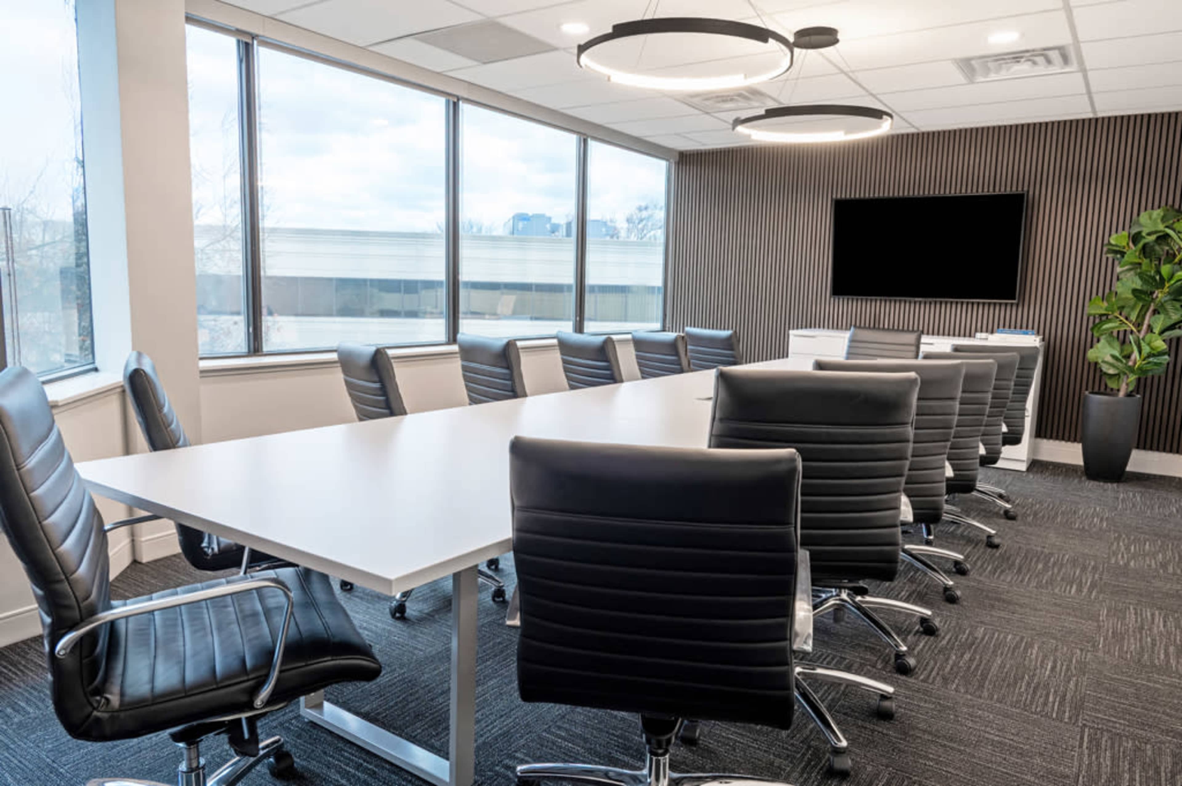 The image shows a spacious conference room with a long white table, multiple black leather chairs, large windows, and a wall-mounted screen.