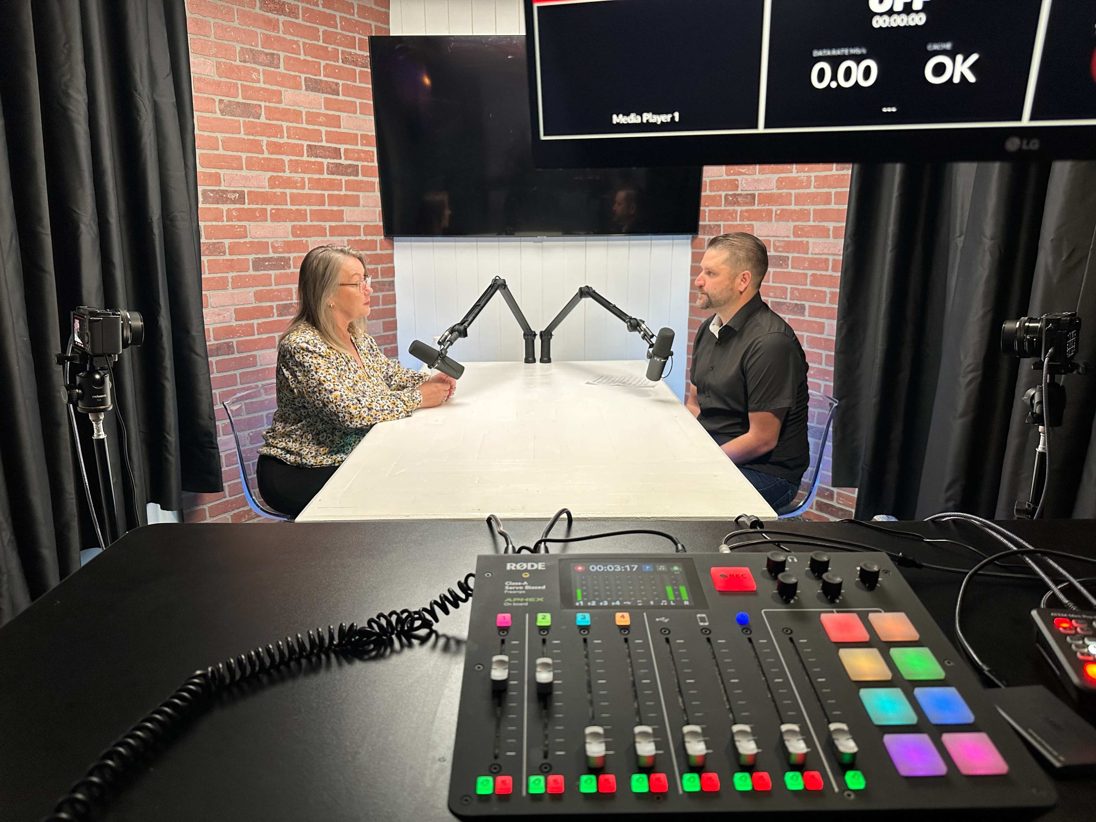A woman and a man sit at a table with microphones in a studio setting featuring brick walls and a large screen.