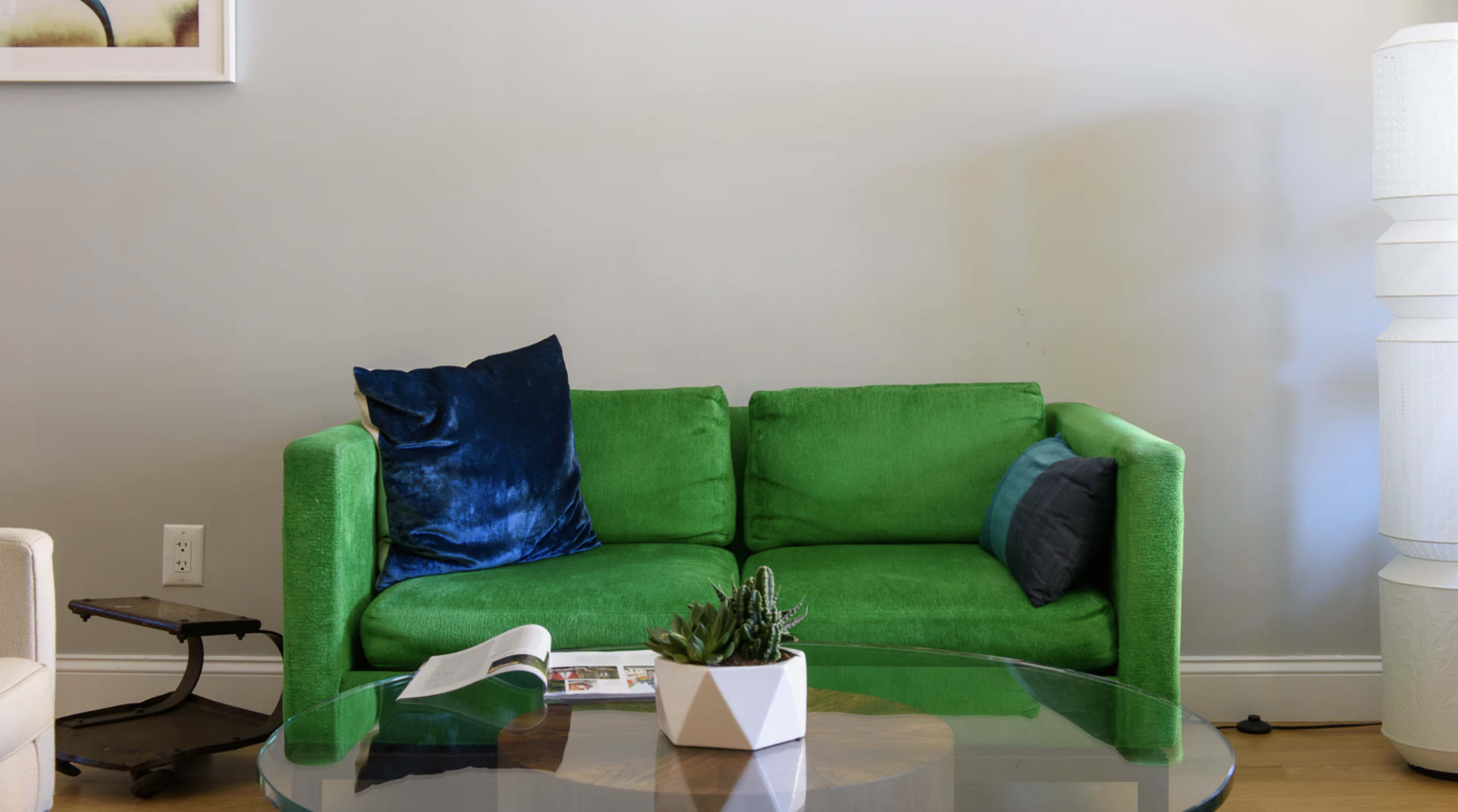 A green sofa with decorative cushions sits in a room beside a coffee table holding a magazine and a small plant.