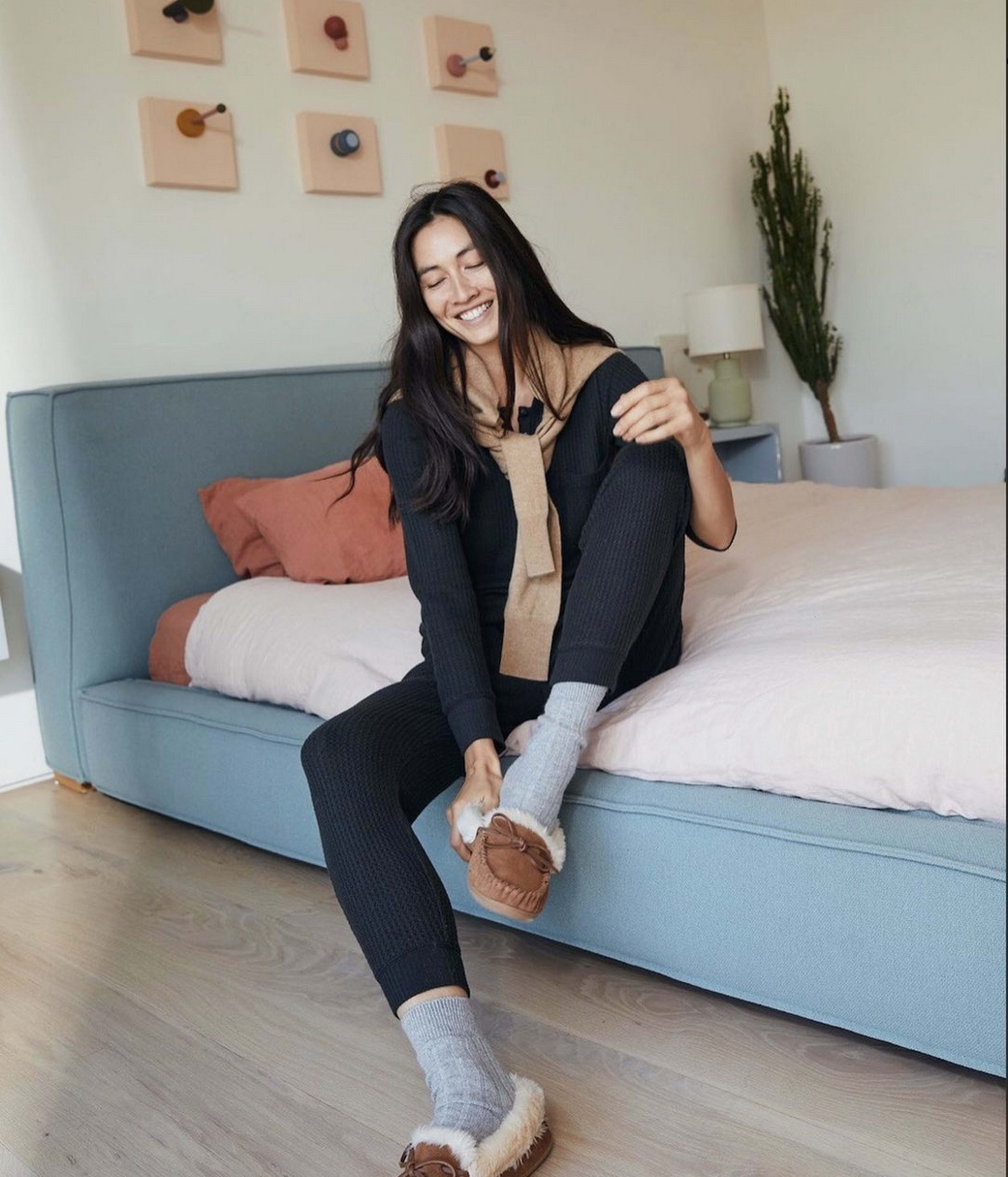 A woman sits on a bed wearing loungewear and slippers, smiling in a well-lit room with decorative wall art.