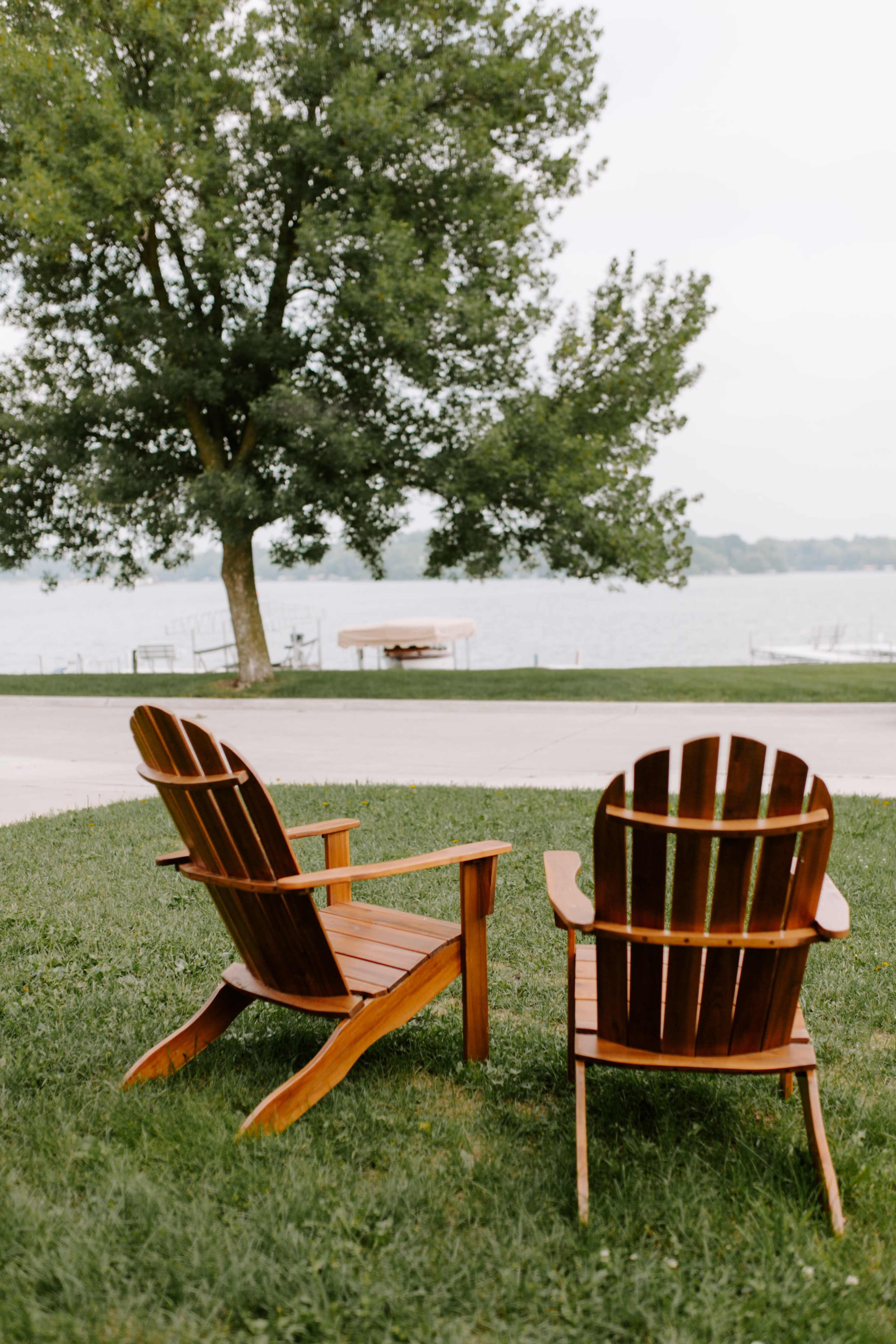 Two wooden Adirondack chairs are placed on grassy land overlooking a calm lake.