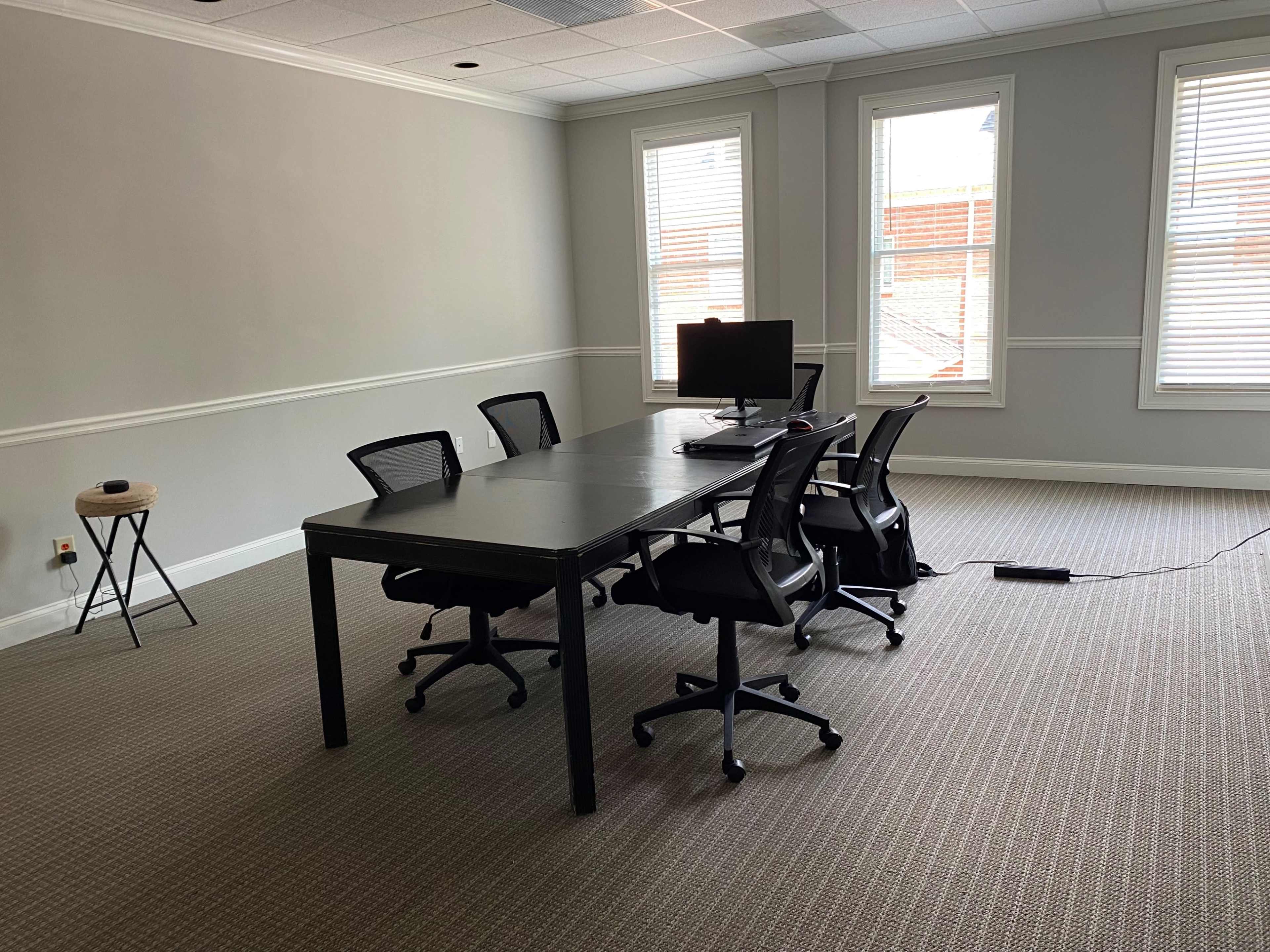 A spacious meeting room features a long black table surrounded by several chairs and a computer monitor on a stand.