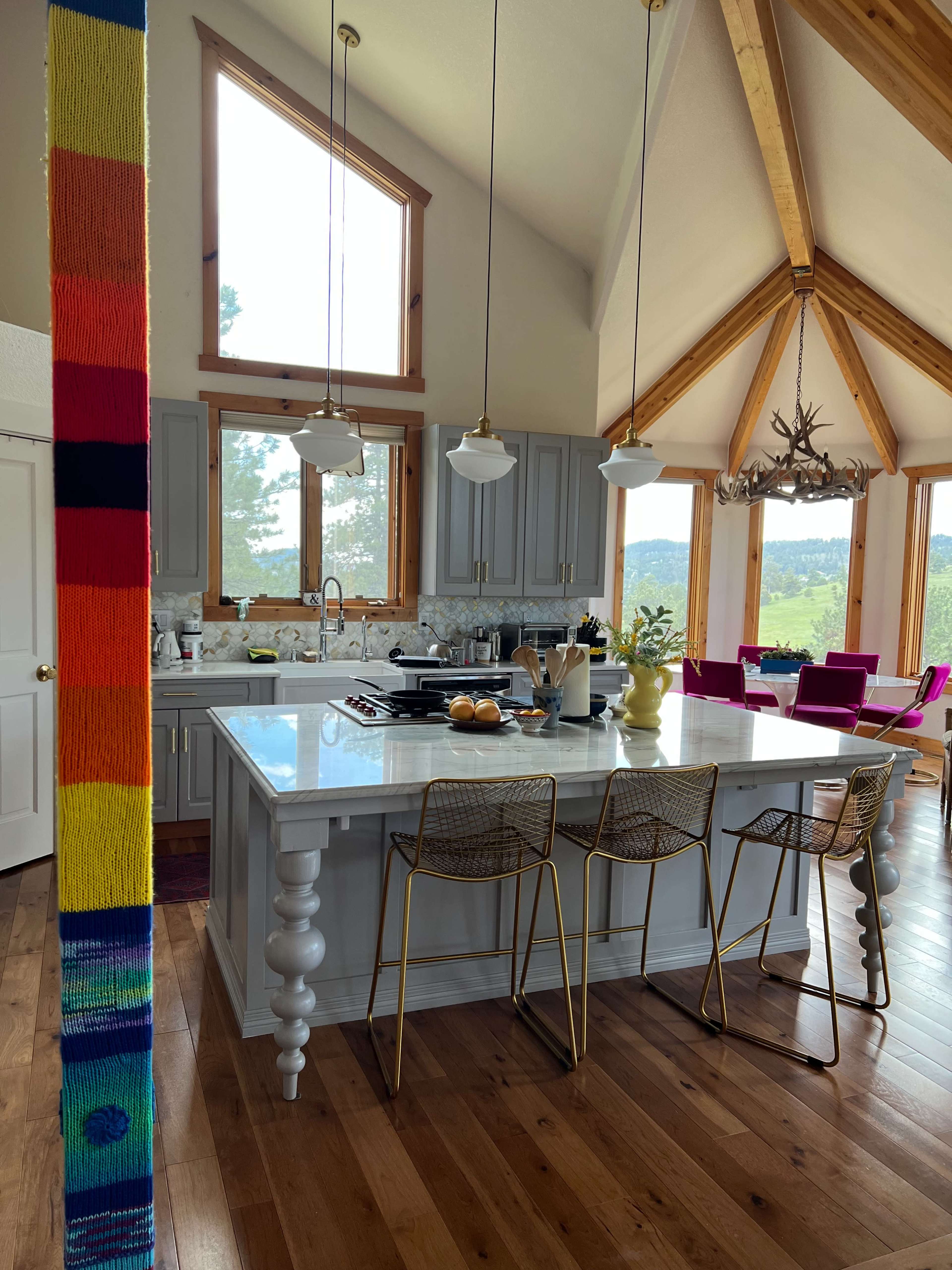 The image shows a modern kitchen with a large island, three bar stools, and a view of greenery through the windows.