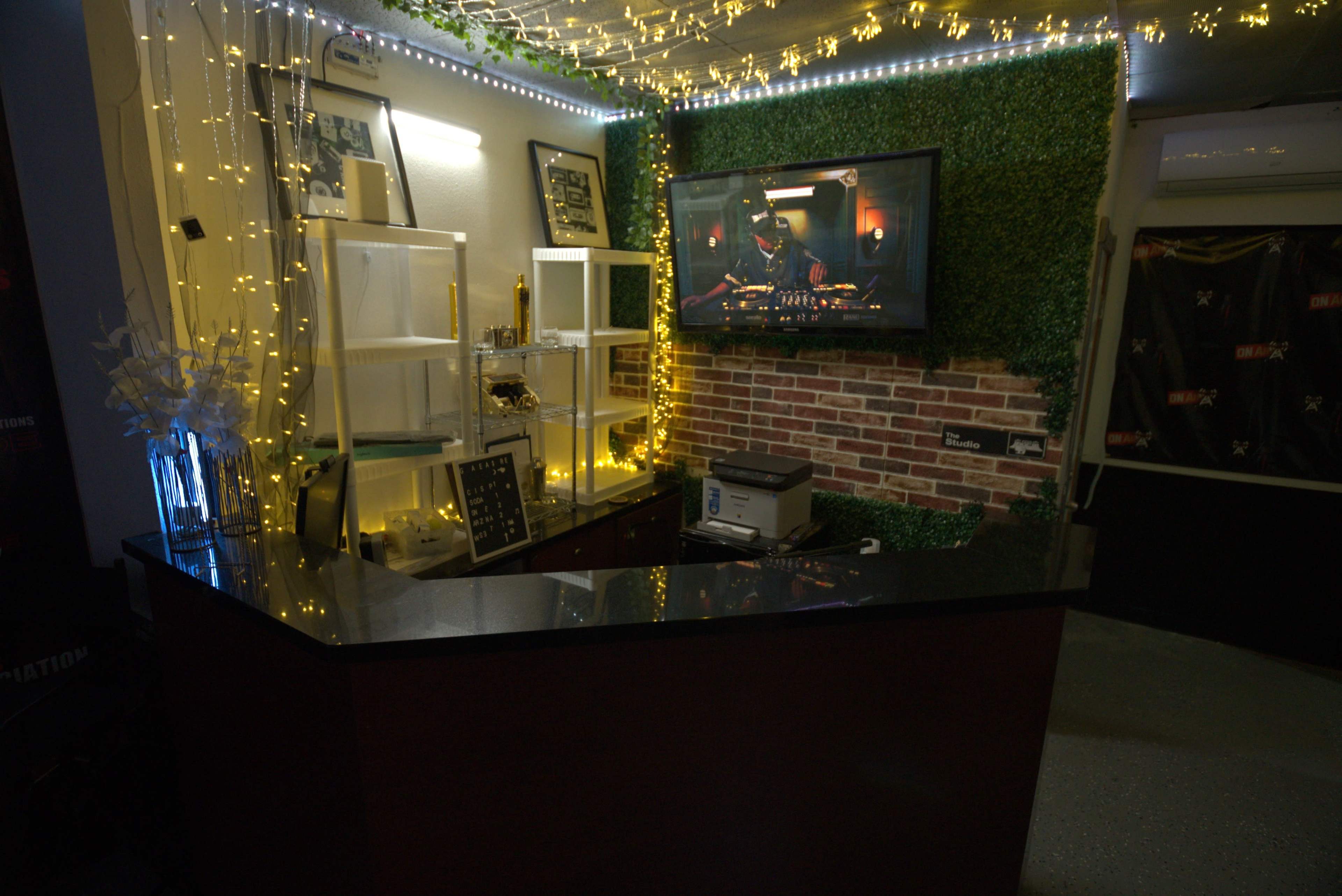 The image shows a reception area with a dark counter, shelves with decorative lights, a television on the wall displaying a chef, and a brick accent wall.
