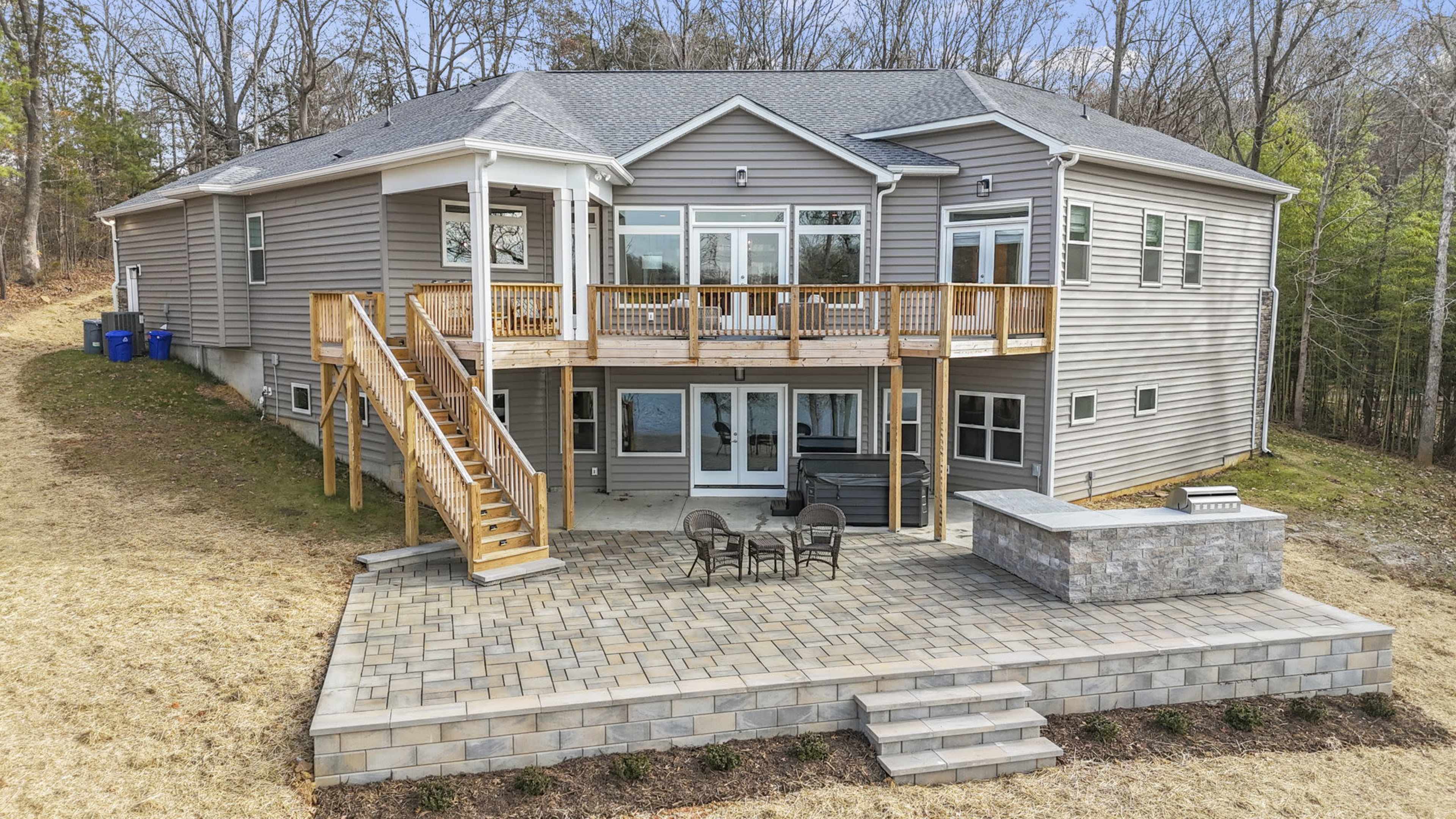 The image depicts a large gray house with multiple windows and a wooden deck, accompanied by stone stairs and a patio area in the backyard.