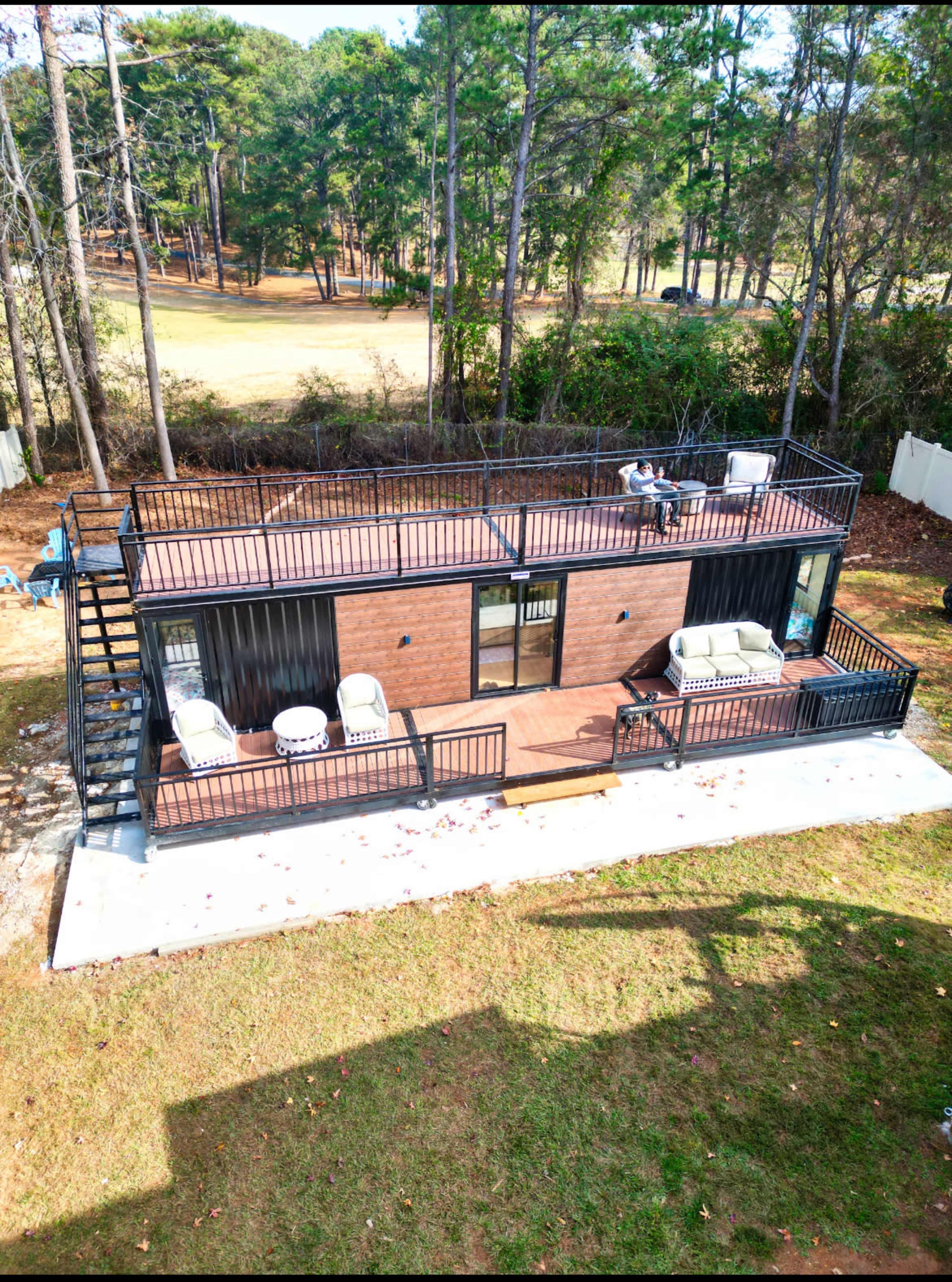 A two-story modern building with a rooftop terrace, surrounded by trees and a grassy area.