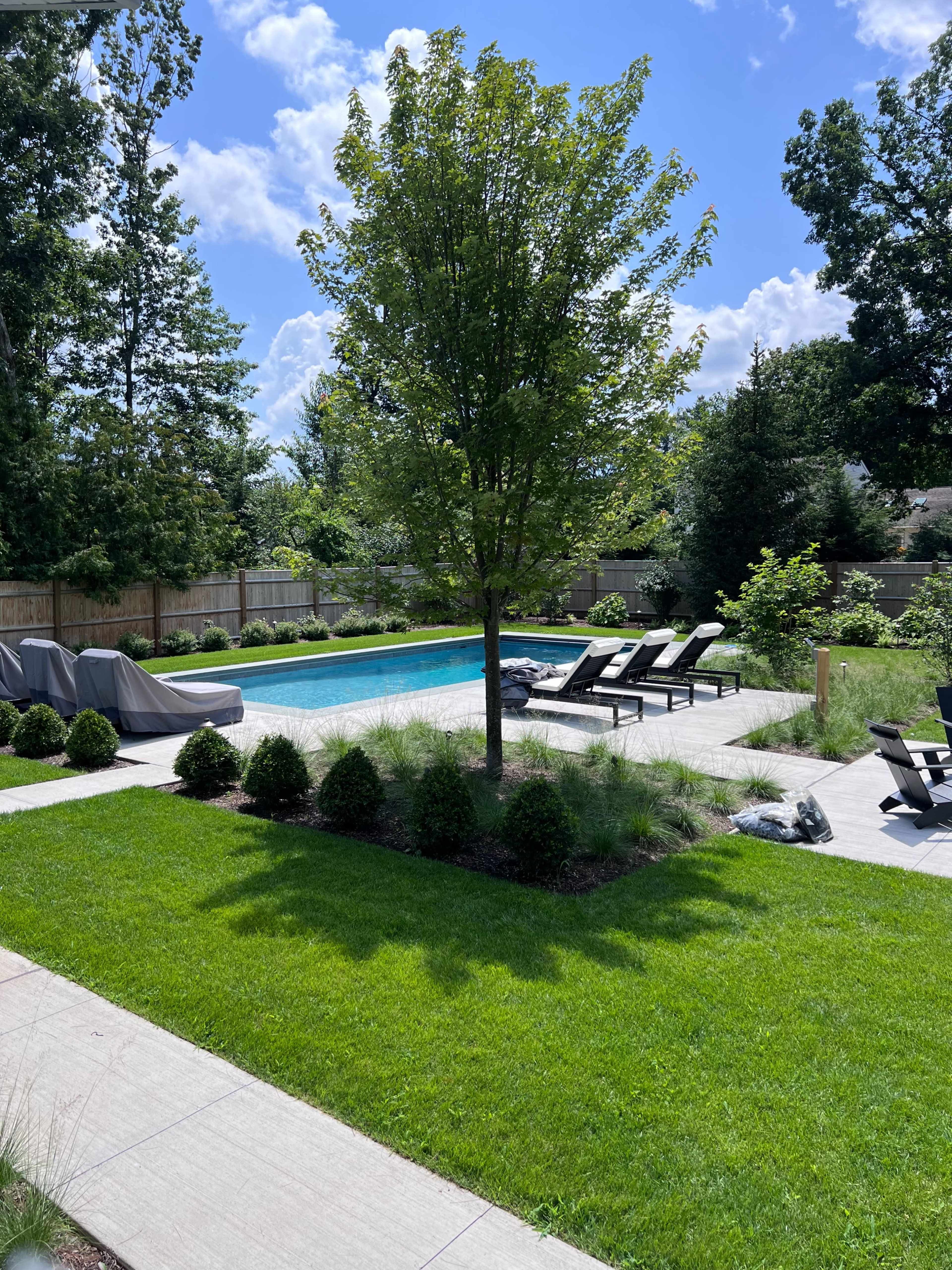 The image shows a backyard with a swimming pool surrounded by lounge chairs, neatly manicured grass, and trees in a sunlit setting.